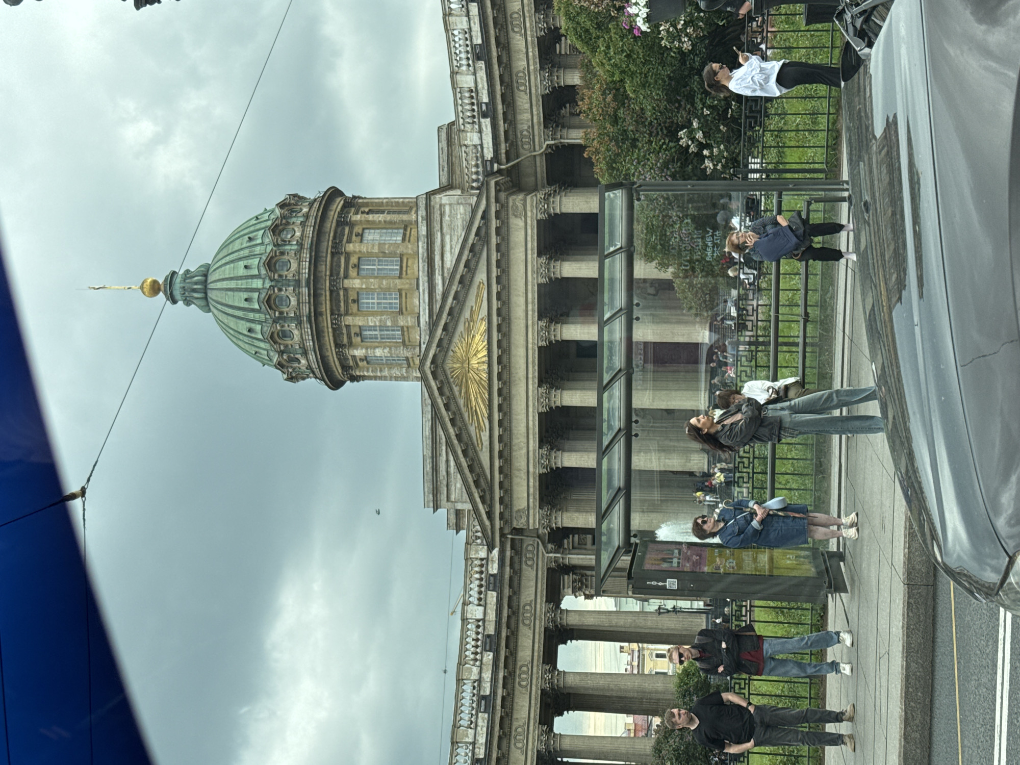 In Saint Petersburg, Russia, a group of people are seen at a bus stop in front of the Kazan Cathedral, a prominent landmark. The sky is overcast with a cloudy appearance.
The Kazan Cathedral, with its distinctive green dome topped by a golden spire, is a grand structure featuring classical architecture with numerous columns and arches. The facade is a pale yellow color.
At the bus stop, there are several individuals waiting. On the left, two men are standing. One, on the far left, wears a black t-shirt and grey pants. Next to him, a man in a red jacket and blue jeans stands with sunglasses on. To the right of them, a woman in a denim dress and white sneakers holds a light blue umbrella. Further to the right, a woman in a dark jacket and jeans stands with her arms crossed. Another woman in a white shirt and black pants is seen looking at her phone, and further back, a woman in a blue and white patterned shirt and black pants sits on a bench, holding a blue backpack. Several other figures are visible in the background, some seated on benches behind a fence.
The bus stop itself is a modern glass and metal structure. In the foreground, the hood and windshield of a dark-colored car are visible, obscuring the lower part of the scene. The ground appears to be a sidewalk and a road. Lush green bushes and trees are present behind the bus stop and fence, with some white and pink flowers blooming. The overall atmosphere is that of a cloudy day in a busy urban environment.
