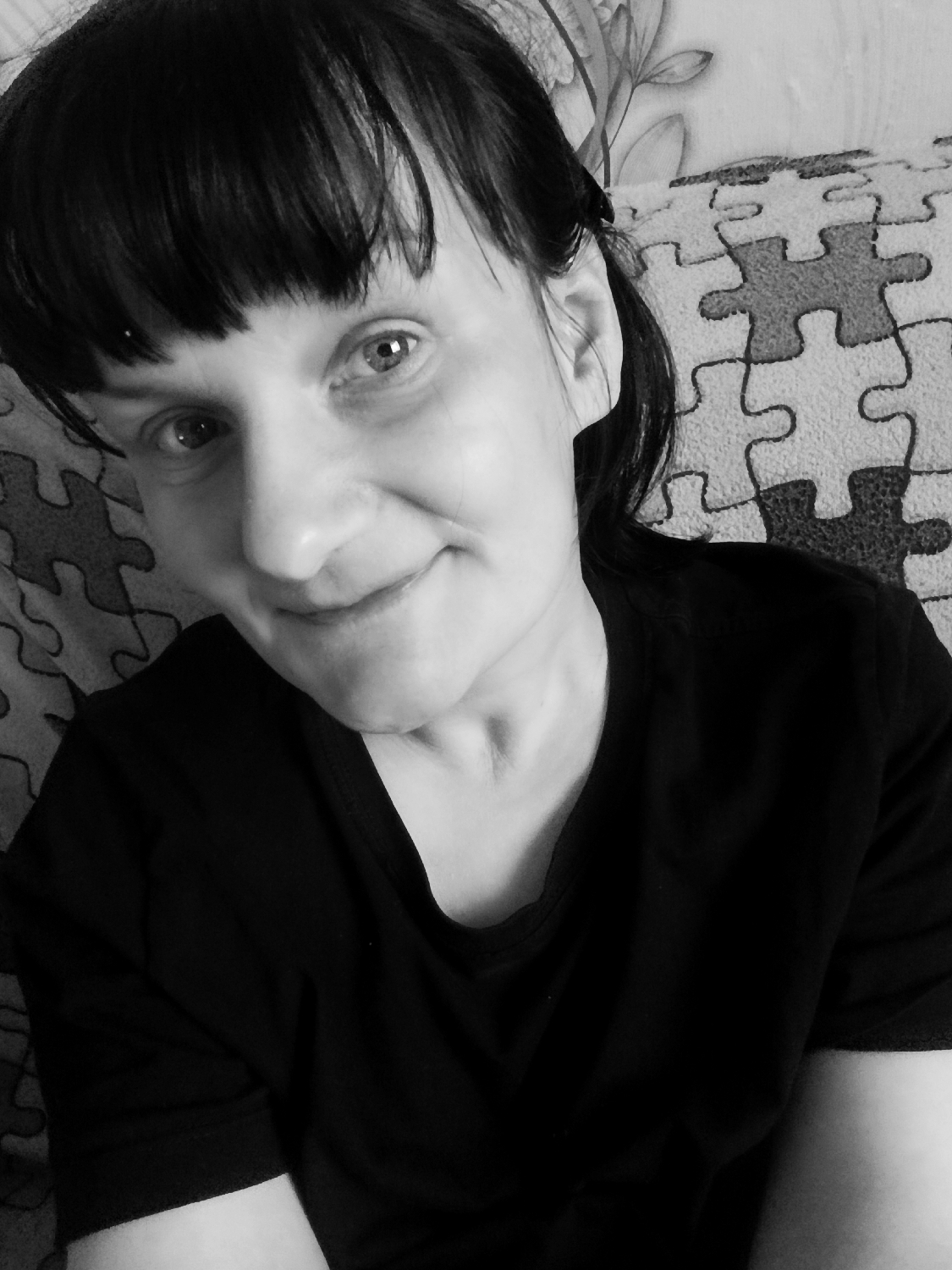 This black and white medium shot captures a woman looking directly towards the viewer with a gentle, slight smile. She has dark hair styled with bangs covering her forehead, and the rest of her hair is pulled back, partially visible behind her ears. Her eyes are open and attentive, with subtle highlights. She appears to be resting or sitting comfortably.
The woman is wearing a plain, dark (likely black) t-shirt with a rounded neckline. The fabric shows subtle folds and shadows, indicating a soft material. Her shoulders and upper arms are visible in the lower part of the frame.
The background behind her is composed of two distinct patterns. To her right and behind her back, there is a prominent design resembling interconnected puzzle pieces, arranged in a grid-like fashion with alternating shades of grey. To her left, behind her head and shoulder, a softer, more organic pattern with floral or leafy motifs is visible, possibly on a wall hanging, curtain, or upholstered furniture. The overall setting suggests an indoor, domestic environment, likely a home in Фоки, Russia.
The woman's expression conveys a sense of calm and a friendly demeanor. The lighting appears soft and diffused, creating gentle shadows. No text is visible within the frame. The photograph is likely a selfie given the angle and closeness of the shot.