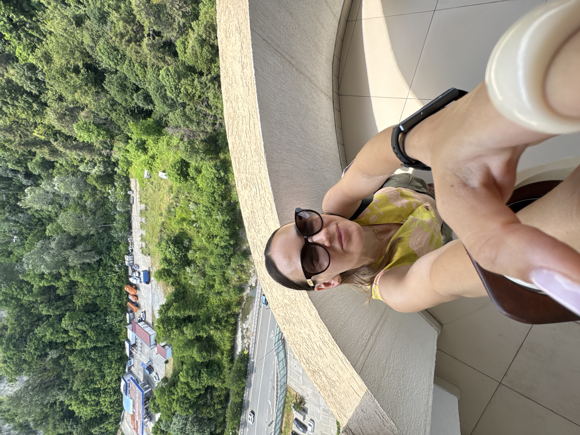 A woman is taking a selfie on a balcony overlooking a lush, green landscape. The woman is wearing sunglasses and a yellow and pink floral top. She has a fitness tracker on her left wrist and is wearing long, light-colored fingernails. The balcony has a textured, beige railing and a tiled floor that is partially illuminated by sunlight, suggesting it is daytime.
Below the balcony, a dense forest covers a hillside. A road runs alongside the forest, with several vehicles visible, including cars, trucks, and orange utility vehicles parked near some buildings. The scene appears to be from a slightly elevated vantage point, offering a broad view of the natural surroundings and the urban elements nestled within it. The lighting suggests a bright, possibly sunny day. The overall impression is one of a scenic, outdoor location, likely a resort or residential area in a place with abundant greenery, consistent with a location like Sochi, Russia. The woman's expression is relaxed, and she appears to be enjoying the view and the moment.