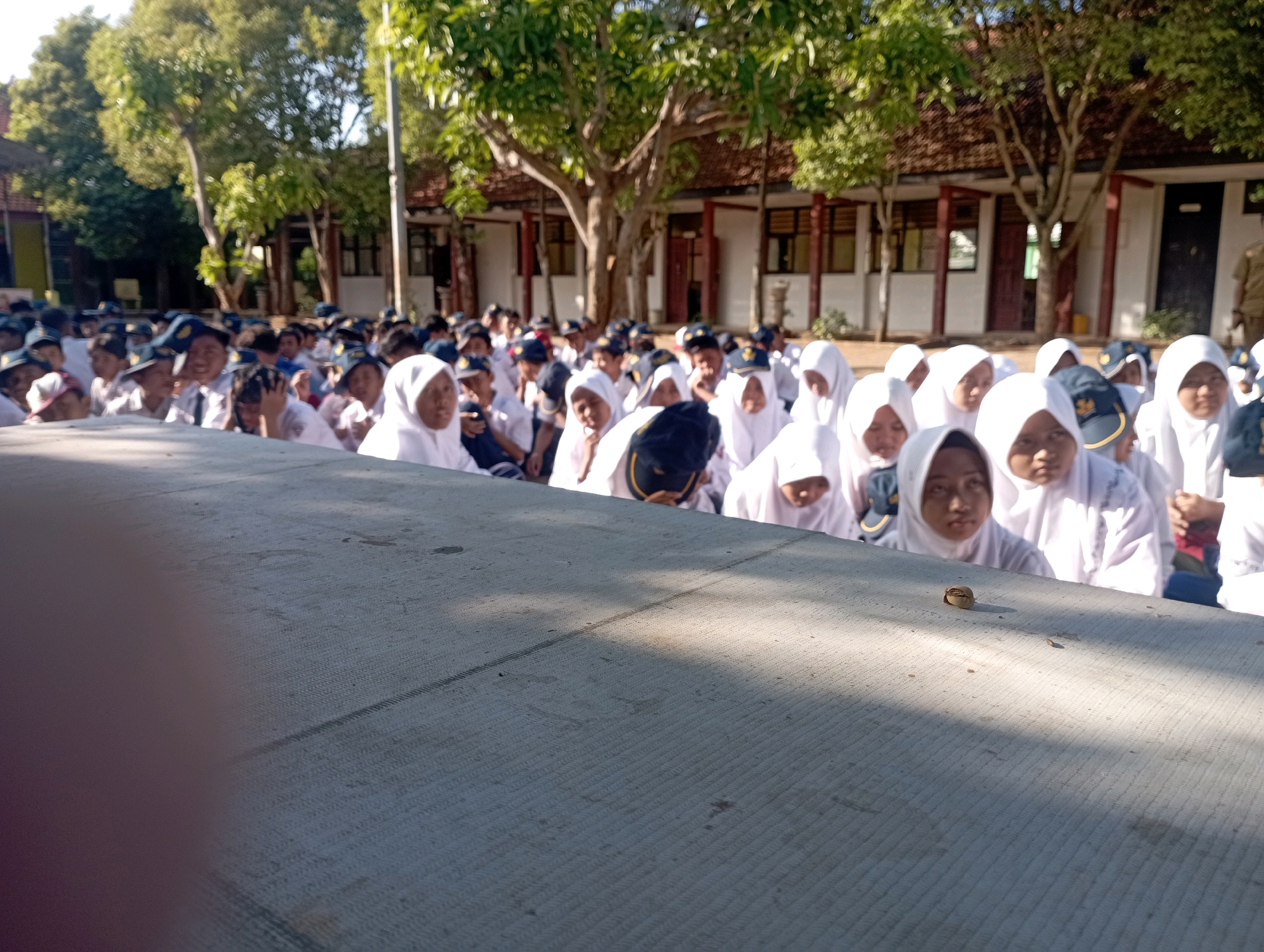 In Banyuurip, Indonesia, a group of approximately 30 to 40 students, predominantly girls wearing white hijabs and uniforms, are assembled outdoors in front of a building with red tile roofing. The students are seated or standing in rows, facing forward, with some visible in the foreground and others further back. The setting appears to be a school or educational institution, indicated by the uniform attire of the students and the architecture of the building. The ground in the foreground is a concrete surface, partially shaded by dappled sunlight filtering through trees. The overall atmosphere suggests an organized gathering or event.
