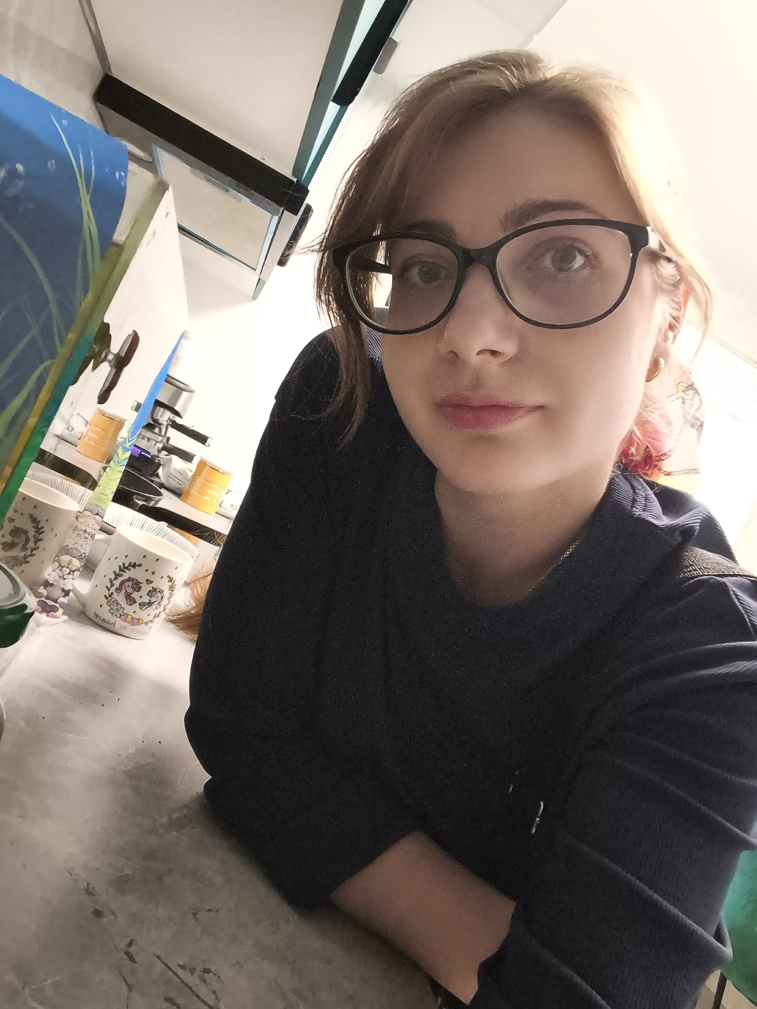 In a medium-angle selfie, a young woman with light brown hair, wearing black-rimmed glasses and a dark blue long-sleeved shirt, leans forward and looks directly at the camera. Her hair is styled in a bob with bangs. She has light makeup on, with a hint of pink on her lips and cheeks. A delicate gold hoop earring is visible on her right ear.
The setting appears to be indoors, possibly a kitchen or a room with shelves. In the background, a coffee maker and several colorful cylindrical containers, likely coffee or sugar containers, are visible on a counter. Two mugs with whimsical designs, possibly featuring gnomes and unicorns, are also present. To the left, a decorative panel with blue and green abstract art is partially visible. The lighting suggests it might be daytime, with natural light possibly coming from a window to the right, casting a warm glow on the woman's face. The overall mood is casual and relaxed. No visible text is discernable in the image. The location is identified as Podkletnoe, Russia.