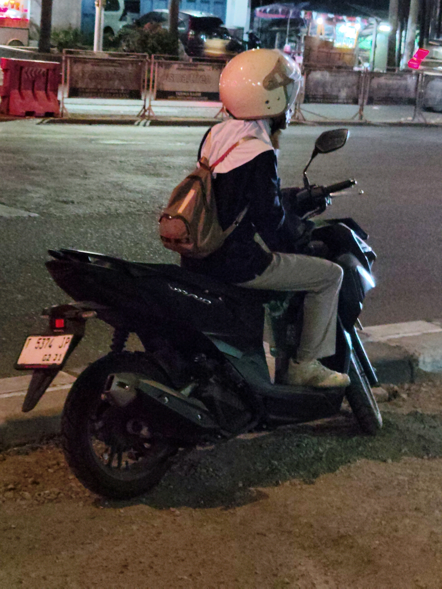A person wearing a light beige helmet and a dark blue jacket with white accents is seated on a dark colored scooter at night. The person has a light brown backpack slung over their shoulders. The scooter has a license plate with the characters "T 5374 JP 02 21". The scene appears to be on a paved street in an urban area. In the background, there are streetlights and what appear to be stalls or shops. The overall lighting suggests it is nighttime. The location is identified as Purwakarta, Indonesia.