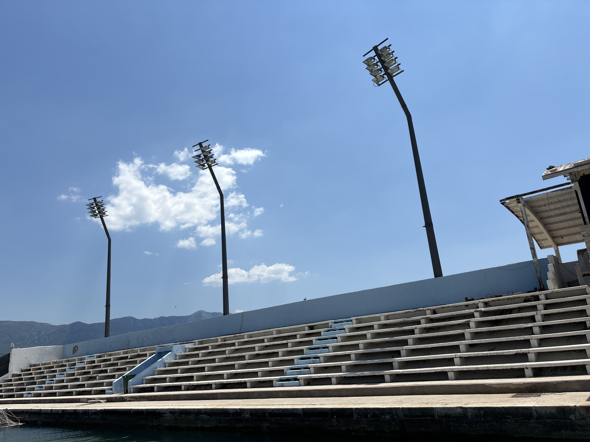 The image depicts the outdoor seating area of a sports venue, likely a stadium or amphitheater, located in Budva, Montenegro. The seating consists of tiered concrete bleachers, appearing somewhat weathered. Three tall light poles, each equipped with multiple spotlights, are prominently visible against a clear blue sky with scattered white clouds. In the background, a range of mountains can be seen. Part of a structure, possibly a roofed seating area or maintenance building, is visible to the right. The foreground shows the edge of the water, suggesting the venue is situated near a harbor or the sea. No people are present in the image.