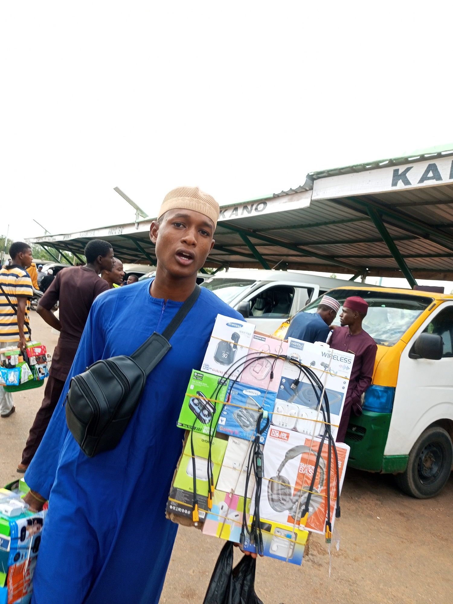The image depicts a street scene in an outdoor market, possibly a transportation hub given the presence of vehicles. A young man in a blue tunic and a light brown cap is prominently featured in the foreground, holding a large stack of merchandise, primarily electronics accessories like headphones and chargers, bound together with string. He is wearing a black fanny pack slung across his chest. In the background, several other people are visible, some carrying baskets of goods, and a yellow and white vehicle with a green stripe is parked. Above, a corrugated metal roof structure with the word "KANO" visible suggests a location in Kano, Nigeria. The overall atmosphere appears to be bustling and commercial.