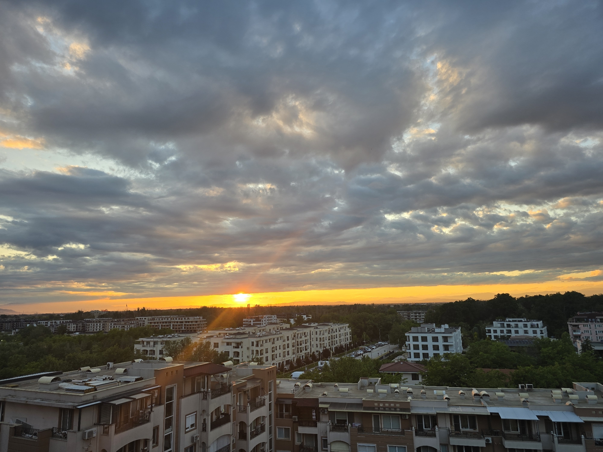 The image shows an urban landscape at sunset or sunrise. Predominantly, there are buildings of varying sizes and architectural styles, suggesting a residential area in the city of Unknown, Unknown. The structures include what appear to be apartment complexes or multi-story residences, some with balconies and tiled roofs. Green trees are interspersed among the buildings, hinting at some degree of green space within the urban setting.
The sky is filled with dramatic cloud formations illuminated by the sun, with the sun appearing on the horizon creating a vivid orange glow. The clouds range from darker, more ominous gray to lighter, brighter hues where the sun's light penetrates. The lighting suggests that it might either be sunset or sunrise, but the overall illumination suggests sunset. There are no people visible. The buildings are illuminated by the sun, creating shadows and highlighting the architectural details.