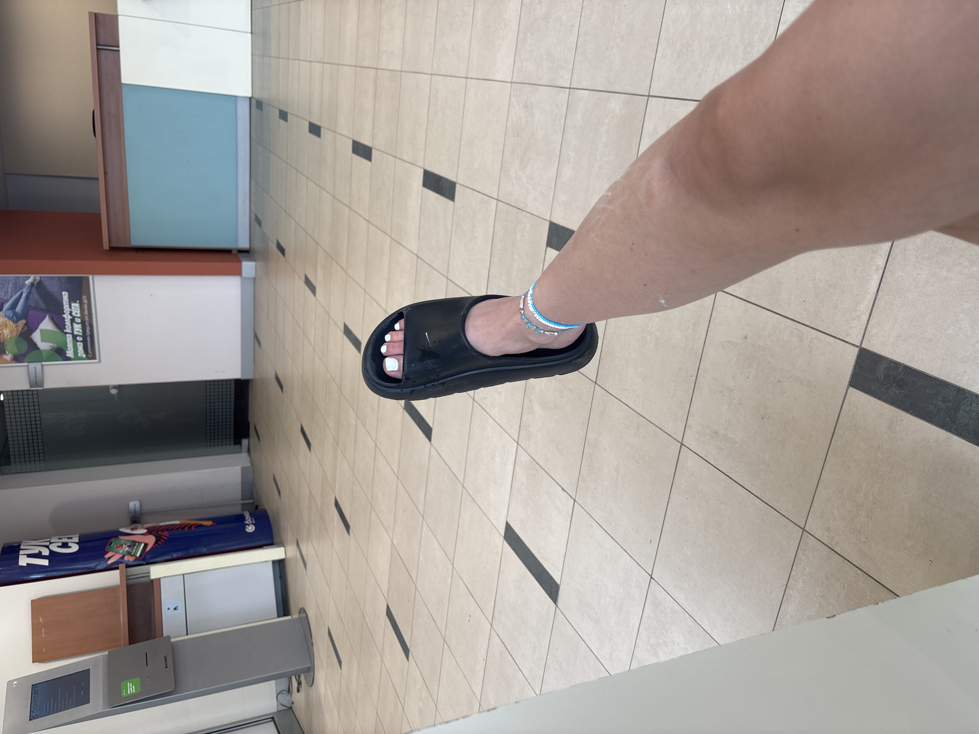 The image captures a first-person, downward view of a person's leg and foot in an indoor public setting. The foot wears a plain black slide sandal, and the toenails are painted white. Two delicate anklets, one blue and one white/light blue beaded, adorn the ankle. The skin on the leg appears pale with some faint, lighter marks.
The floor is composed of light beige square tiles with regularly spaced black rectangular accent tiles, indicating a commercial or institutional environment. In the background, various architectural and commercial elements are visible. On the left side, there's a light brown counter or desk, behind which are light blue and white panels. Below the counter, a poster is partially visible on a white wall. The poster contains Cyrillic text, reading "МЕЖДУНАРОДНА ЗОНА НА ГТК и СБ" and "София-предизвикателство" (Sofia-challenge).
Further along, a prominent blue cylindrical display stand with text and graphics is visible, bearing partially legible Cyrillic text, including "ТУК СЕ..." and the logo for "БАНКА ДСК" (DSK Bank). Adjacent to this display is a grey ATM or information kiosk, which also displays the "БАНКА ДСК" logo on its screen panel. The overall lighting suggests an interior during daytime or a well-lit space.