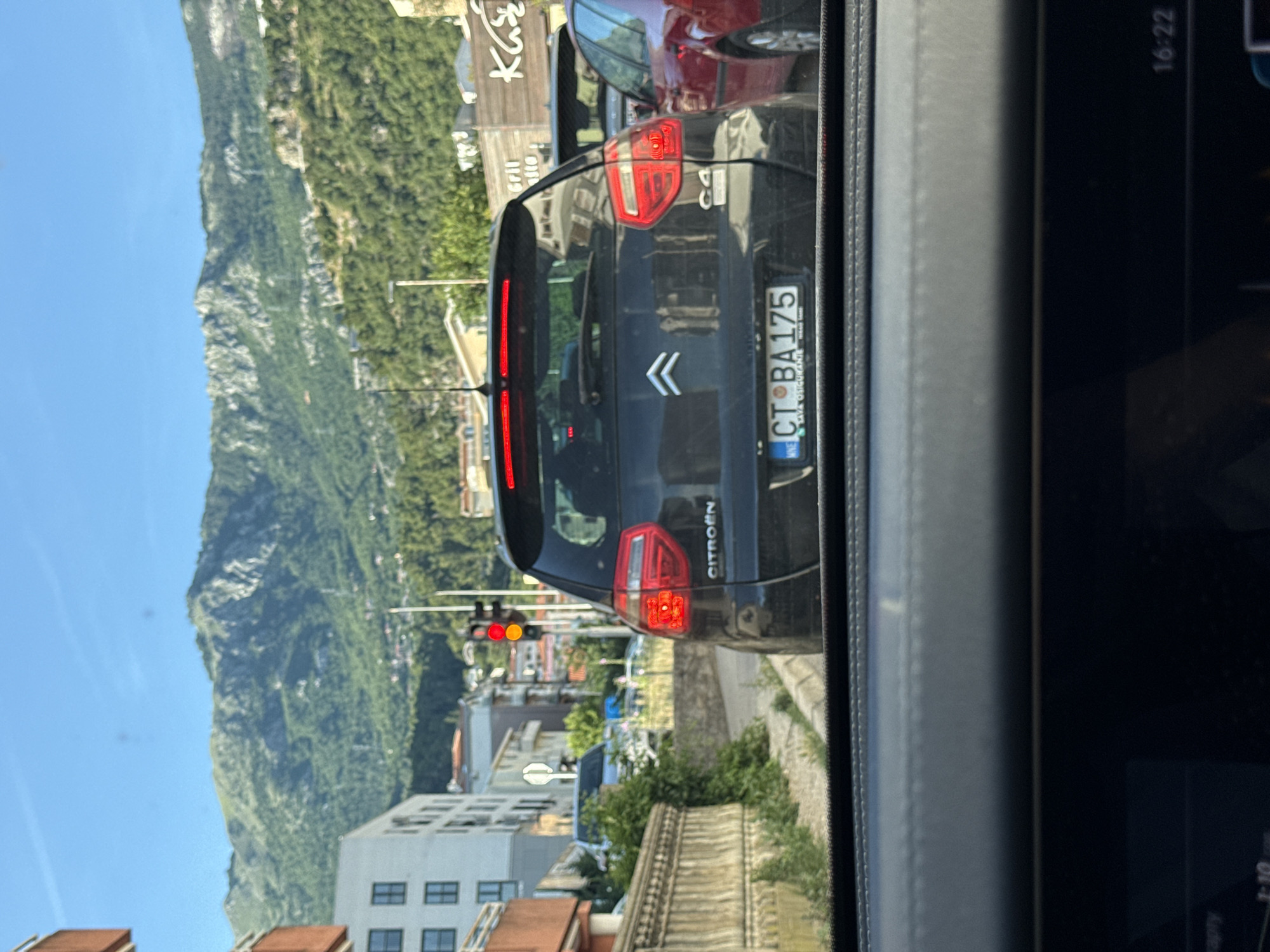 The image depicts a street scene in Budva, Montenegro, likely during daylight hours with a clear blue sky. The foreground is dominated by the interior of a vehicle, showing a dashboard and part of a windshield. Outside the vehicle, a black Citroën C4 is the primary subject, parked in traffic. The car's rear lights are illuminated. Its license plate, CT BA 175, is visible, indicating it is registered in Montenegro.
The car is in a traffic lane, with other vehicles behind it and to its right. A red car is partially visible to the right of the Citroën. The background reveals a city environment with buildings of varying heights and architectural styles. One building on the left appears to be modern with visible window frames. Further in the distance, lush green mountains rise, characteristic of the Budva region. A traffic light is visible, showing red and yellow signals, suggesting a regulated intersection. Signs on buildings in the background are partially obscured but appear to indicate commercial establishments. The overall impression is a snapshot of a typical day in a busy Montenegrin city.