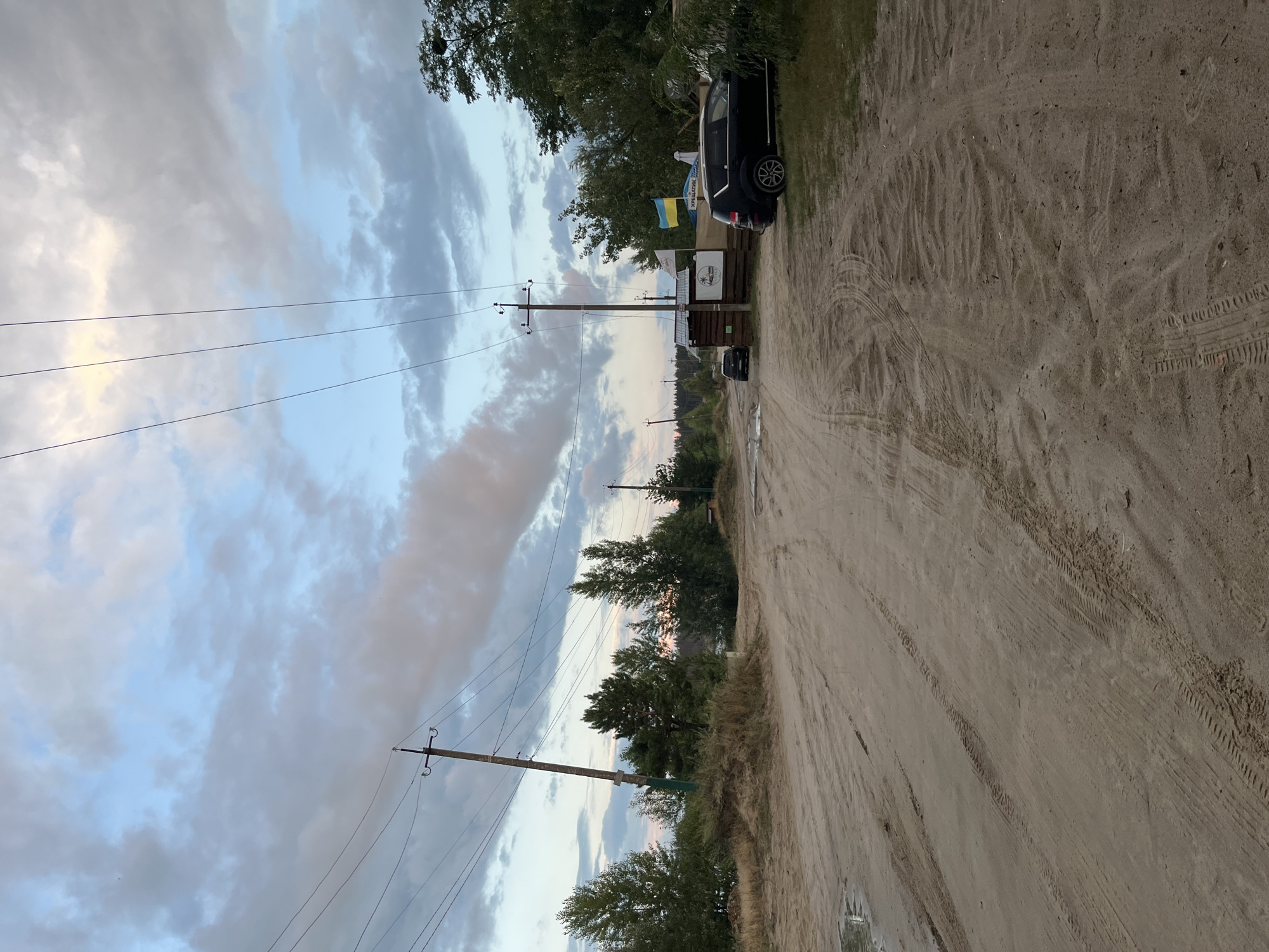 The image depicts a dirt road leading through a wooded area near Bezliudivka, Ukraine. The sky is partly cloudy with hues of blue and gray. Power lines and utility poles are visible overhead. On the right side of the road, a dark SUV is parked next to a small wooden structure. A Ukrainian flag is displayed prominently on the structure. Trees and foliage line the sides of the road, with some sandy areas and puddles of water suggesting recent rain. The road shows tire tracks, indicating vehicle activity.