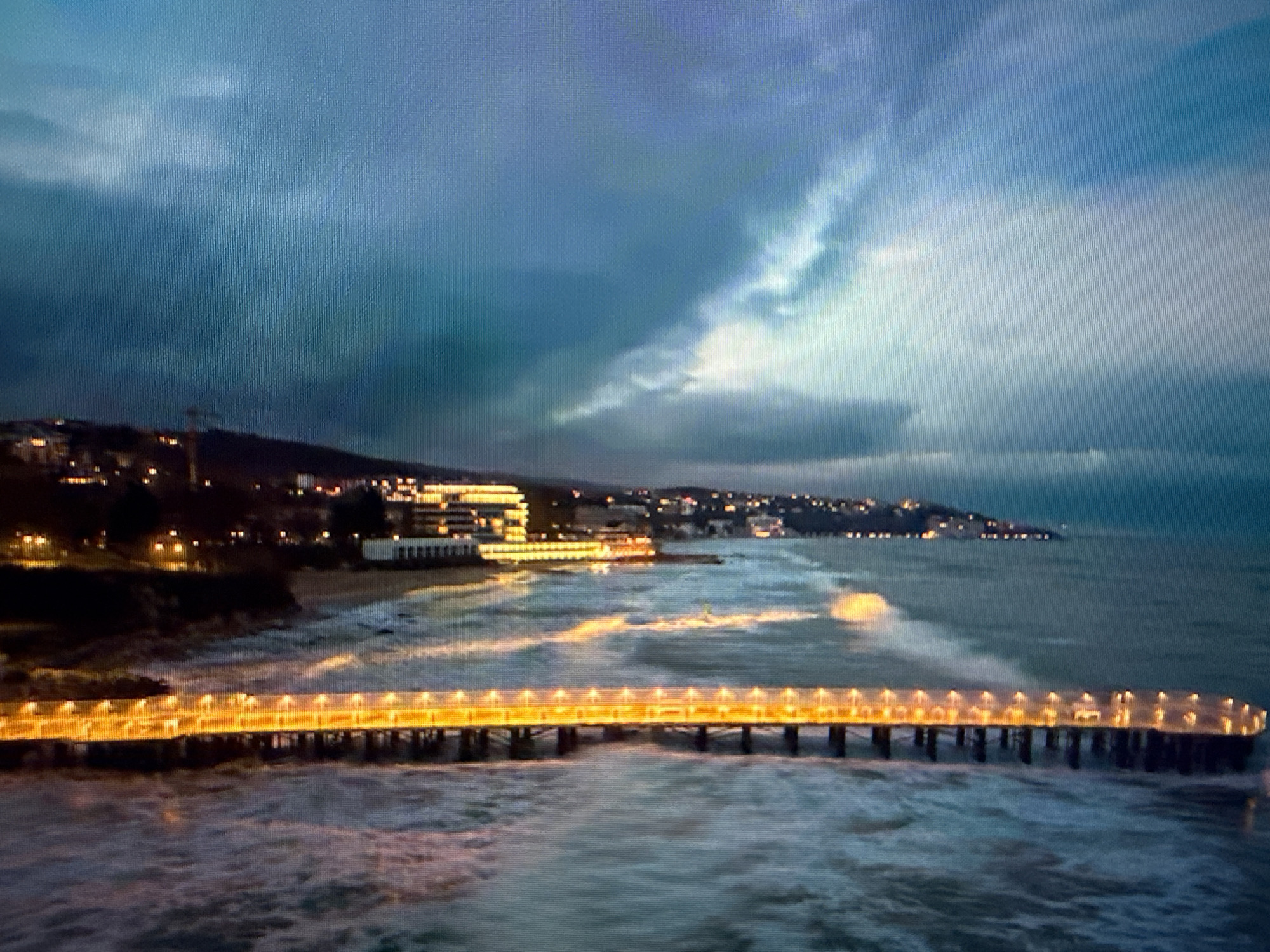 The image shows a pier extending into the ocean at night. The pier is illuminated with warm yellow lights along its length. Waves are breaking against the pier's supports. The coastline is visible in the background with buildings and streetlights dotting the shoreline. The sky is overcast with a dramatic cloud formation. The picture does not show any elements indicating that the location is in Sofia, Bulgaria, which is located far from any sea coast.