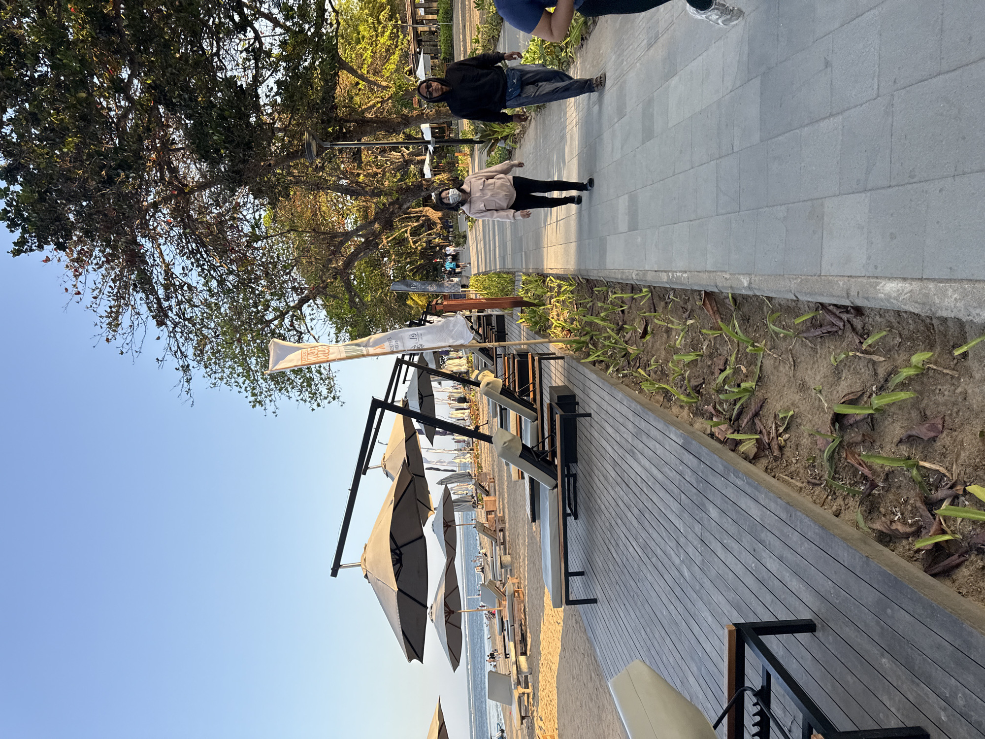 The image shows a beach path in Denpasar, Indonesia, on a sunny day. The path is made of grey pavers and is lined with a planter box containing young green plants. On the left side of the path is a wooden deck with several unoccupied lounge chairs and umbrellas, suggesting a resort or public beach area. Beyond the deck, a sandy beach with more lounge chairs and umbrellas is visible, leading to the ocean where a few people are walking near the water.
On the right side of the path, two individuals are walking away from the camera. The person in front is wearing a light pink jacket and black pants and has a white mask covering their face. The person behind them is wearing a black hoodie and dark pants, with sunglasses and a blue fanny pack. Further down the path, more people are visible walking and strolling under the shade of numerous trees that line the area. The sky is clear and blue, indicating good weather. The overall scene depicts a well-maintained public or resort area designed for relaxation and recreation by the beach.