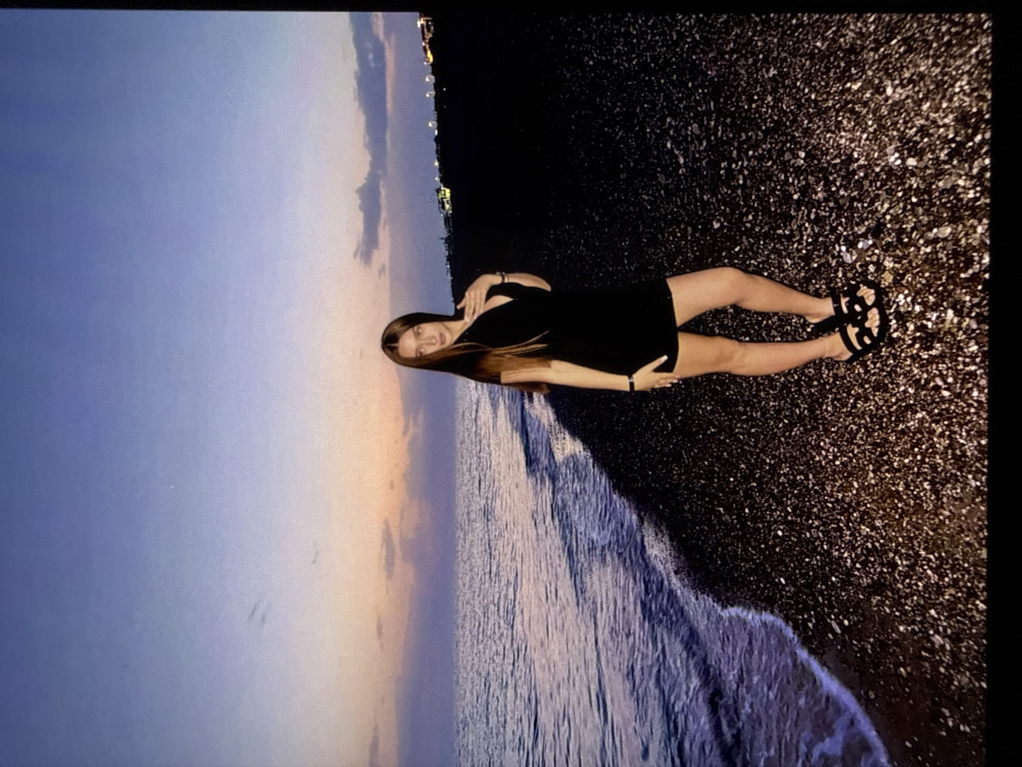 A young woman with long brown hair stands on a pebble beach at dusk. She is wearing a short black dress and black sandals. The sea is to her left, and the sky is a gradient of purple, pink, and blue. In the distance, on the right, are lights from a city. The woman is looking at the camera and has her left hand on her chest. The scene is calm and serene.
