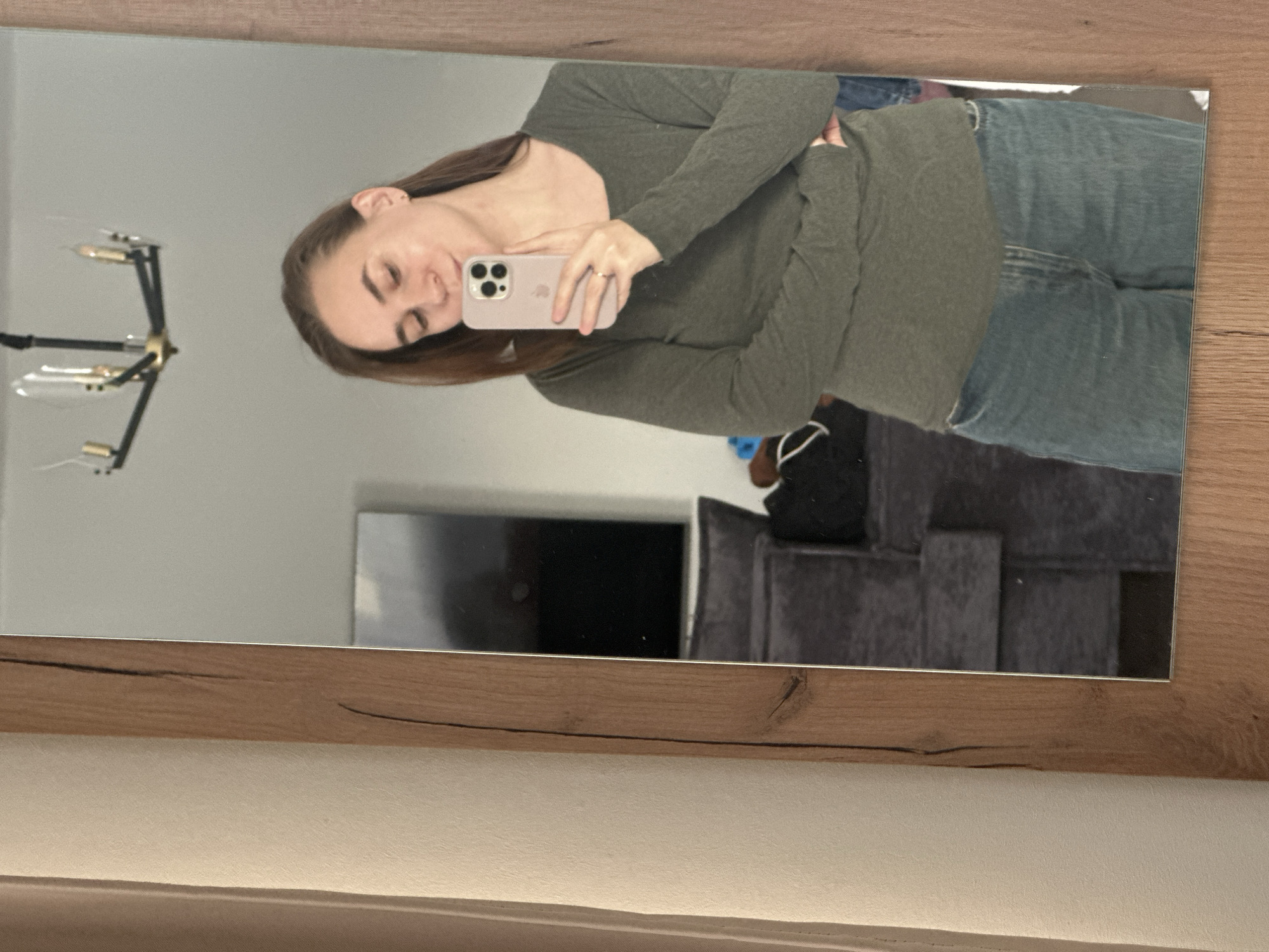 A woman is reflected in a mirror with a wooden frame. She is holding a light pink smartphone in her right hand, presumably taking the photo. She is wearing a long-sleeved olive green shirt and blue jeans. Her brown hair is long and falls past her shoulders.
In the background of the reflection, there's a modern chandelier on the left, a dark television screen, and a grey sofa. The scene appears to be indoors, likely in an apartment or house. There is no visual information in the image to confirm the location as Kharnavy, Russia.
