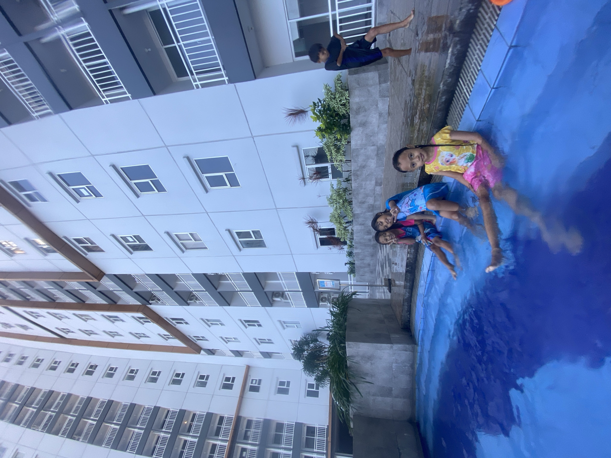 In Sidoarjo, Indonesia, a group of children are enjoying a swimming pool adjacent to a modern, multi-story apartment building. Four young girls are seated on the pool's edge, with their legs submerged in the clear blue water. One girl, wearing a yellow shirt with a cartoon character and pink shorts, smiles at the camera. To her left, another girl in a blue swimsuit and pink shorts also smiles, with her hand raised in a peace sign. Further left, two more girls are visible, one wearing a blue and pink swimsuit and the other mostly obscured. In the background, a young boy in black shorts and a blue shirt stands on the poolside, facing away from the camera and appearing to be in motion. The apartment building, with its white facade and numerous balconies with white railings, dominates the scene. Green foliage and potted plants are arranged around the pool area, adding a touch of nature to the urban setting. The sky is overcast, indicating a possibly humid day.