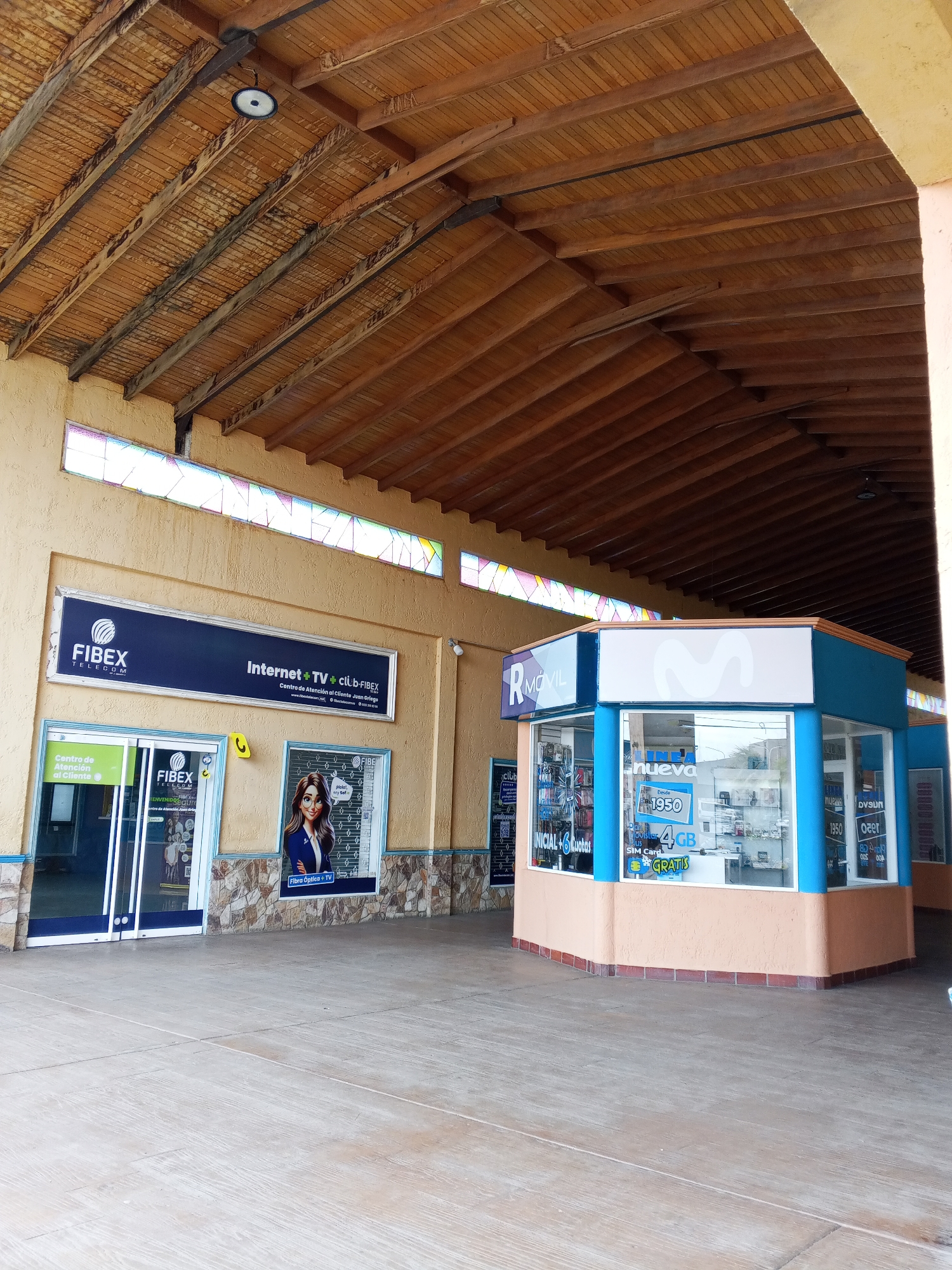 The image captures an outdoor commercial space, likely a shopping arcade, in Juan Griego, Venezuela. The scene is dominated by two storefronts. On the left, a FIBEX Telecom store features a prominent blue sign displaying the company's name and services: "Internet + TV + Club-FIBEX." The store has a glass-door entrance labeled "Centro de Atención al Cliente" and a poster of a woman promoting fiber optics and TV. Adjacent to it, on the right, there is an R Móvil booth, a smaller kiosk-style structure. The booth is painted in blue and peach colors with promotional advertisements, including the price "Desde 1950" and "4GB GRATIS" along with mobile phone displays. The floor is a light-colored, smooth concrete surface. The building's structure features a high, slanted wooden roof with visible wooden beams. There are decorative stained-glass windows lining the top of the building façade. Overall, the image presents a commercial area with telecom and mobile phone businesses.