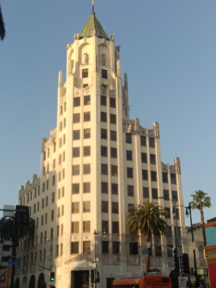 The image depicts the Eastern Columbia Building, located at 849 S Broadway, Los Angeles, California, United States. The prominent subject is a tall, cream-colored Art Deco building featuring multiple stories, rectangular windows, and a distinctive green-pitched roof spire. The building's upper facades are adorned with decorative architectural elements, including sculpted figures and crenellated motifs.
The sky overhead is clear and light blue, indicating daylight. Natural sunlight illuminates the left and front surfaces of the building, casting some areas into shadow on its right side. At street level, several palm trees are visible, along with traffic light poles bearing signal heads and street signs; one sign is partially legible as "Broadway." The rear portion of an orange transit vehicle, likely a bus, is visible at the bottom right of the frame. Parts of adjacent commercial structures with colorful facades are also discernible on the right side of the image. No individuals are visibly present, and no specific actions or interactions are occurring within the scene.