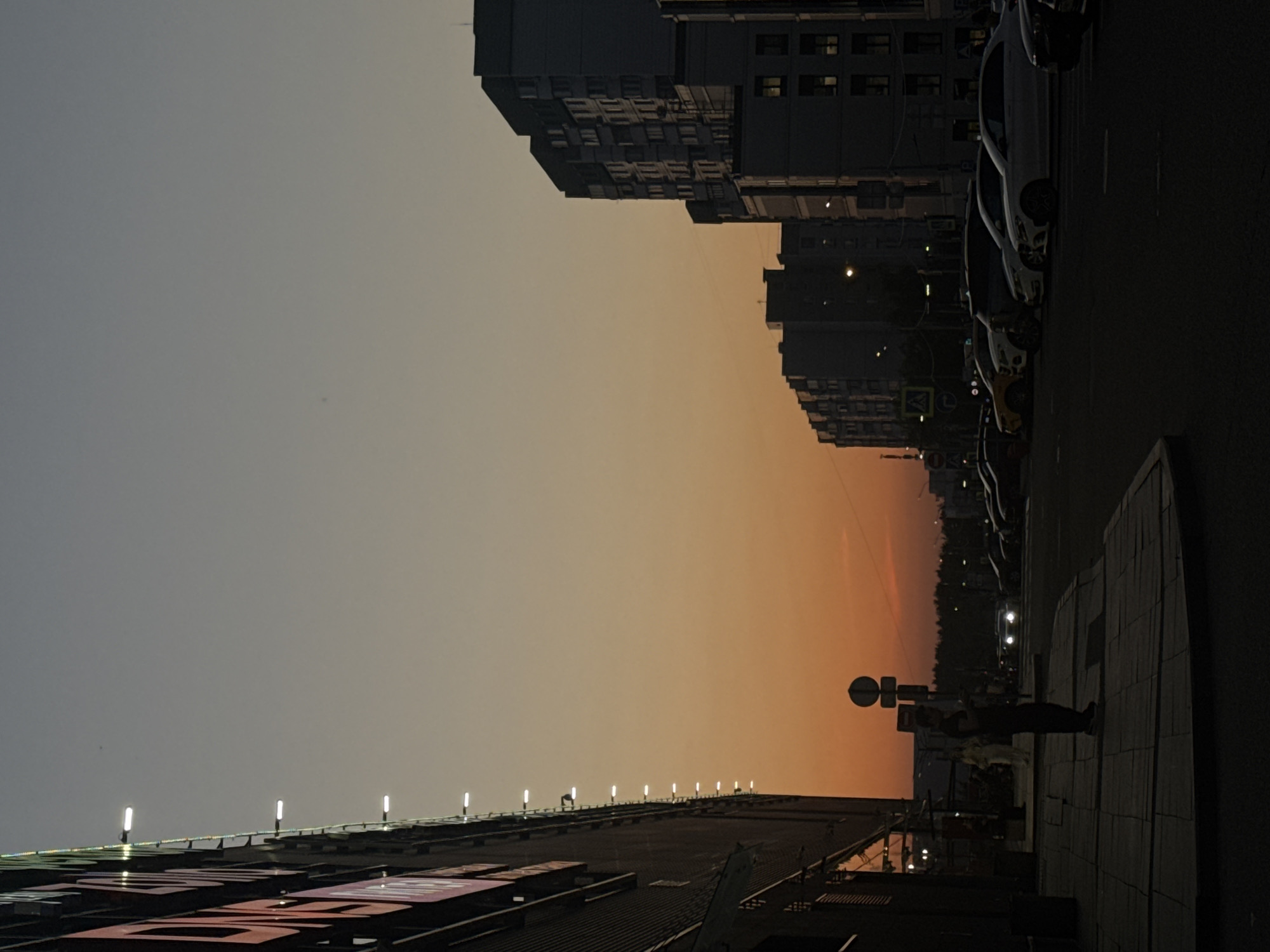 The image shows a street scene in Moscow, Russia at dusk. On the right, cars are parked along a dark road next to a sidewalk with a standing person. A street sign is visible near the person. Tall, dark buildings line the street. The sky transitions from grey to orange. On the left, the edge of a building is lit by a series of lights.