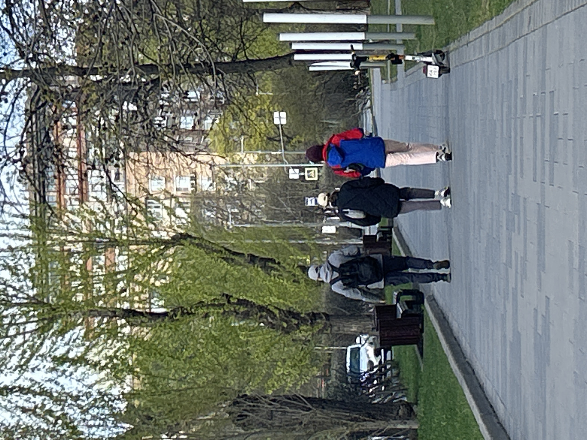 The image captures a daytime scene on a paved pathway in what appears to be an urban park or boulevard in Moscow, Russia, during early spring. The photograph is rotated 90 degrees clockwise.
Three individuals are walking along the pathway from the background towards the foreground. The person closest to the viewer is wearing a dark hoodie or jacket with a backpack and dark pants. Ahead of them is another person in a dark jacket and dark pants, also with a backpack. Further down the path, a third person is visible, wearing a bright red jacket, a blue backpack, and light-colored pants.
The pathway is made of light grey paving stones. On either side, there are strips of green grass and numerous trees. Some trees still have bare branches, while others are beginning to sprout fresh, light green leaves, indicative of early spring. Several park benches are placed along the path. In the background, multi-story buildings with numerous windows are visible through the tree line, suggesting a residential or commercial area. Various streetlights or poles and square road signs are also scattered along the path. A white electric scooter is parked near a cluster of white vertical poles; faint red numbers, possibly "242" or "247," are visible on a small box attached to it. A white vehicle, possibly a car or van, is partially visible further down the path.
The lighting suggests an overcast or slightly hazy day. The overall activity is a casual stroll or commute through a public urban space. No specific event is depicted beyond everyday pedestrian movement.