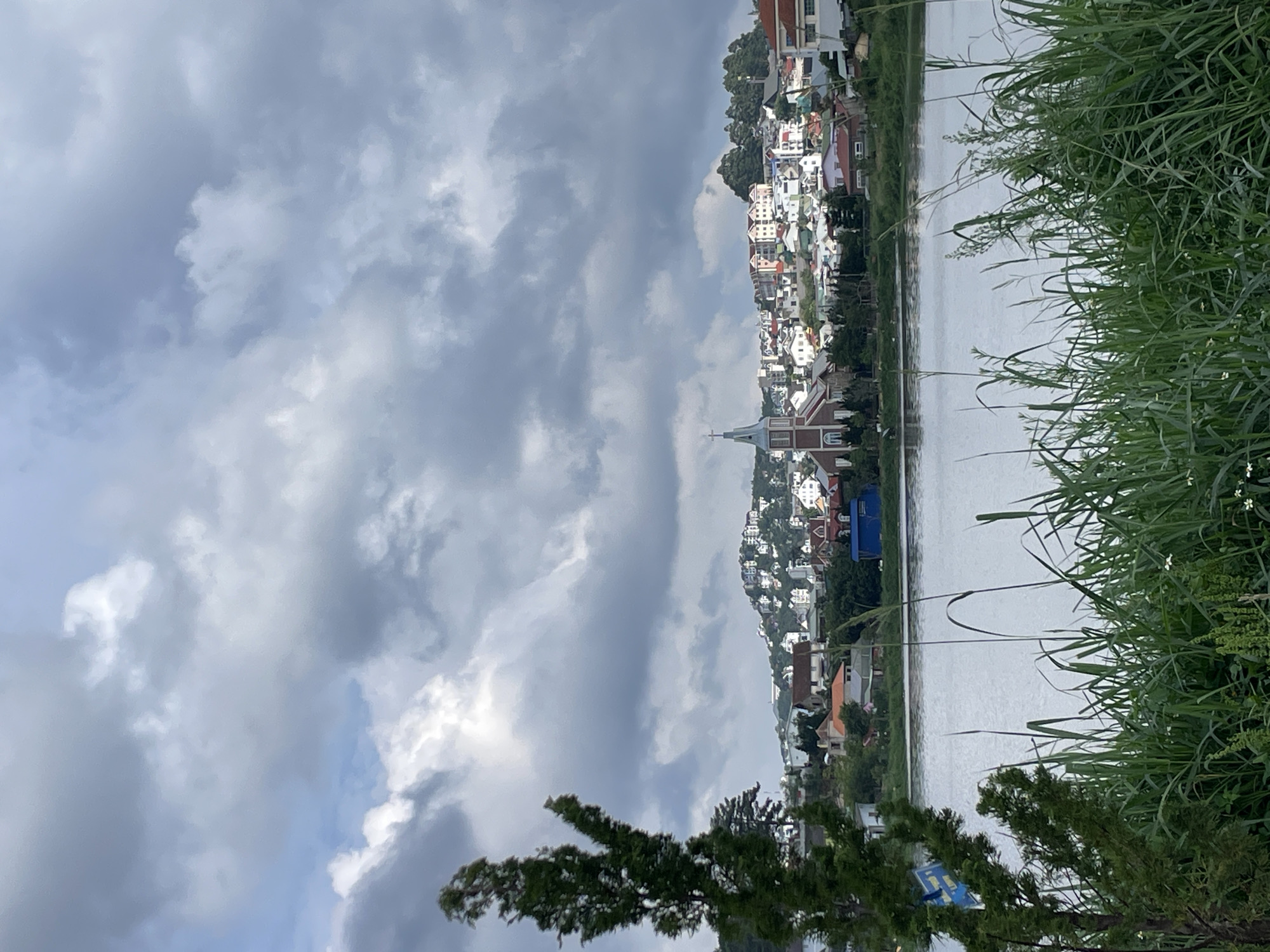 This landscape image, likely taken in Lâm Viên, Đà Lạt, Vietnam, captures a serene view of a hillside town situated beside a body of water under an overcast sky. The horizon line is defined by the calm, greyish water, which stretches horizontally across the middle ground.
On the far side of the water, a densely built town ascends a gentle hill. Numerous multi-story houses with varied roof colors (red, orange, brown, white) dot the hillside, characteristic of the architecture often found in Đà Lạt. A prominent white church with a tall spire and a cross stands out among the buildings, its dark-tiled roof providing a contrast. The town appears nestled within lush greenery, with trees interspersed among the houses.
The sky occupies a significant portion of the upper frame, filled with soft, diffused light filtering through a blanket of grey and white clouds. There are no clear patches of blue sky, suggesting an overcast day.
In the foreground, primarily on the right side, a thick band of vibrant green grass and reeds rises, partially framing the water and the distant town. Some taller stalks of grass reach into the mid-frame. On the lower left and center, dark green tree branches with dense foliage extend into the view, further accentuating the natural elements in the foreground. No people are visible, and there is no discernible text on any signs or buildings. The overall impression is one of peaceful, natural beauty with a touch of urban charm.