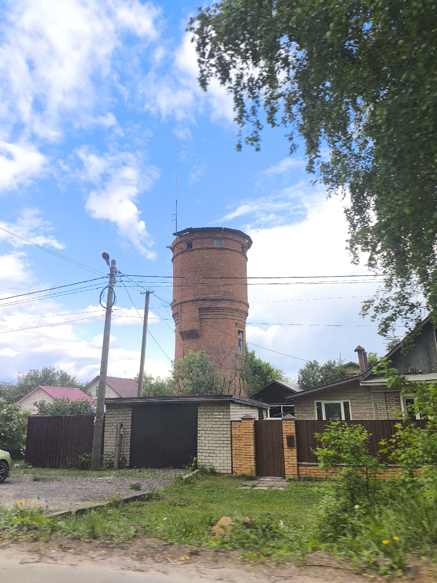 The image depicts a landscape with a tall, cylindrical brick water tower as a central feature. The tower has a weathered appearance, with visible windows and a metal structure on top. Overhead electrical wires extend from a nearby utility pole towards the tower.
In the foreground, there are several houses with different architectural styles and materials, including brick, wood, and metal fencing. The houses are partially obscured by foliage and trees. A paved or unpaved road runs along the bottom edge of the image.
The sky is blue with scattered white clouds. The overall environment appears to be a residential area in a city.