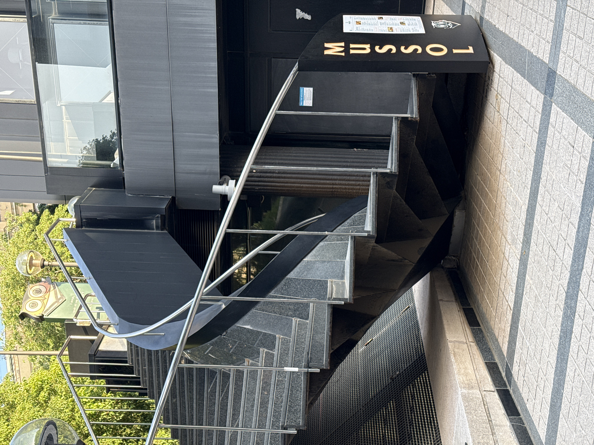 The image depicts the exterior staircase leading to the "Mussol" restaurant in Barcelona, Spain. The staircase is a modern design, constructed with dark grey stone treads and stainless steel railings. The restaurant's signage, with gold lettering, is visible at the top of the stairs. The building adjacent to the staircase has large windows, and greenery is visible in the background. The stairs lead up from a tiled walkway.