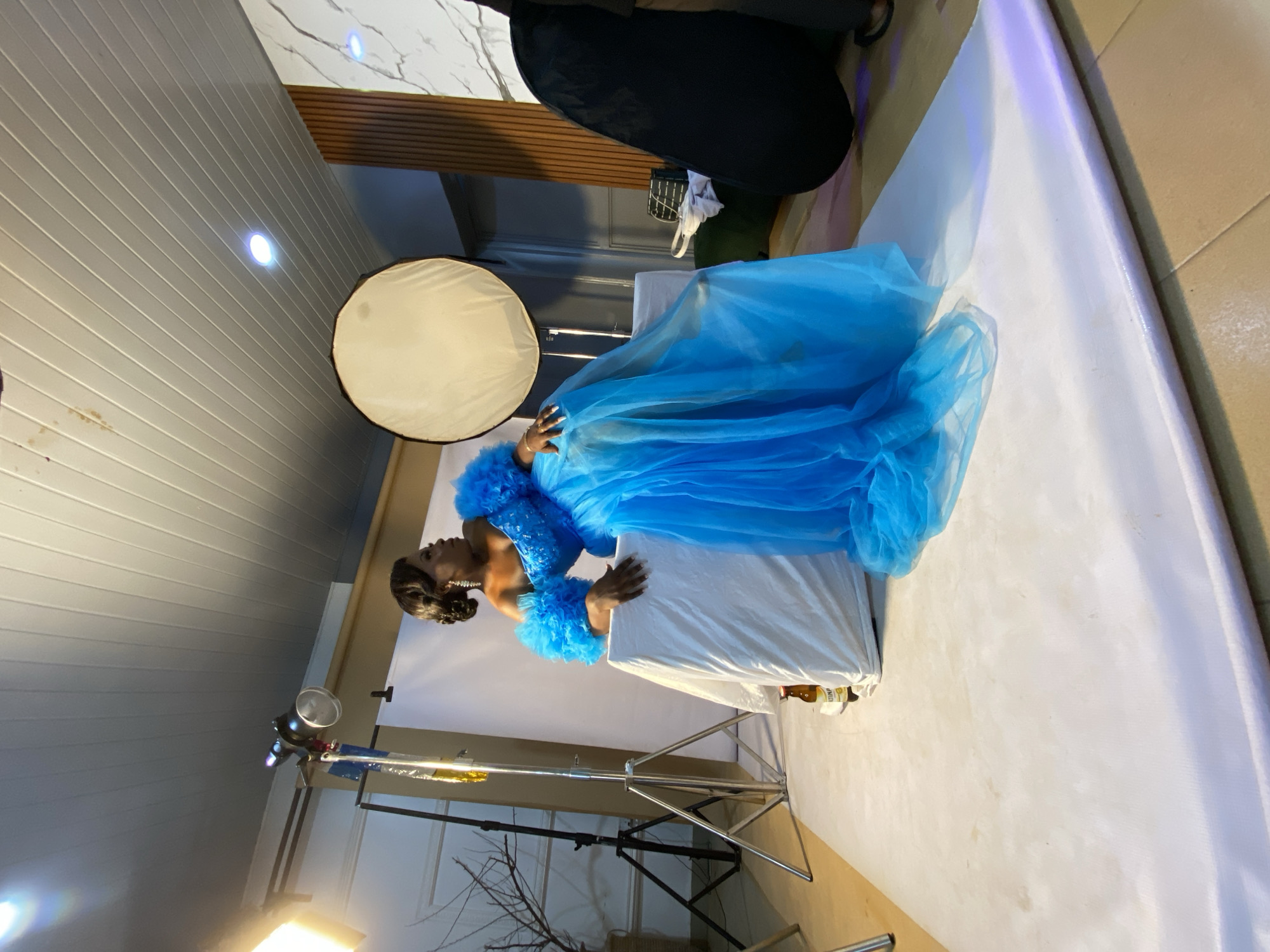 The image depicts a woman posing for a photoshoot in what appears to be a studio setting. She is wearing a voluminous, bright blue tulle gown with ruffled detailing on the shoulders and sleeves. The gown has a sparkly embellishment on the bodice. Her hair is styled in an updo, and she is wearing earrings and a bracelet. The woman is positioned on top of a white covered surface, possibly a table or platform, with her body turned to the side and her head looking upwards. Her hands are resting on the covered surface.
The background features studio equipment, including lighting stands, softboxes, and a large black reflector. The walls are paneled with horizontal planks, and there is a section of marbled wallpaper. The lighting appears to be artificial, consistent with a studio environment. There are no explicit indications of the time of day or weather. The overall impression is a professional or creative photoshoot session. The provided location context is Bukuru, Nigeria, but this is not visually evident from the image itself. No visible text is present in the image.