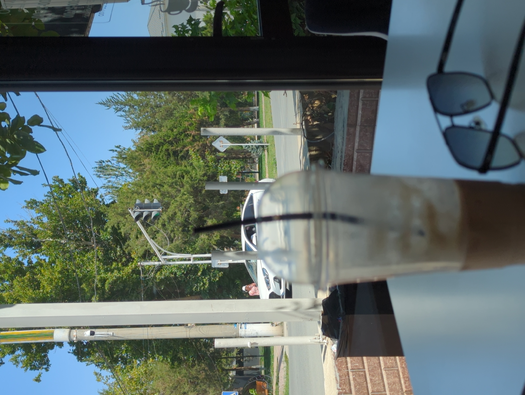 This image captures a view from inside a cafe, looking out onto a street. In the foreground, a blurred plastic cup with a straw and a brown liquid is placed on a white table, alongside a pair of dark-rimmed sunglasses. The exterior scene shows a street with a white car, a traffic light, and a pedestrian wearing a light-colored shirt and a white cap. Trees and poles line the street, and a bright blue sky is visible above. The location is identified as Orto-Boz, Kyrgyzstan.