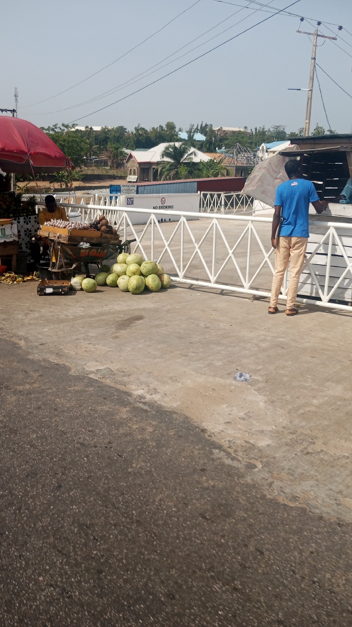 The image shows a scene in Lokoja, Nigeria, on a sunny day. The foreground consists of a road surface transitioning to a concrete pedestrian area. A pile of approximately 15 large, green watermelons sits on the concrete. Next to the watermelons is a small cart with an assortment of fruits and vegetables, attended by a person under a red umbrella. A man in a blue shirt and tan pants stands near a white metal barrier. The background features buildings and foliage under a clear sky. Power lines are visible overhead. A sign on the white barrier reads "OUT" and "NO SMOKING". The general impression is that it is a roadside fruit stand, potentially near a port or customs area.