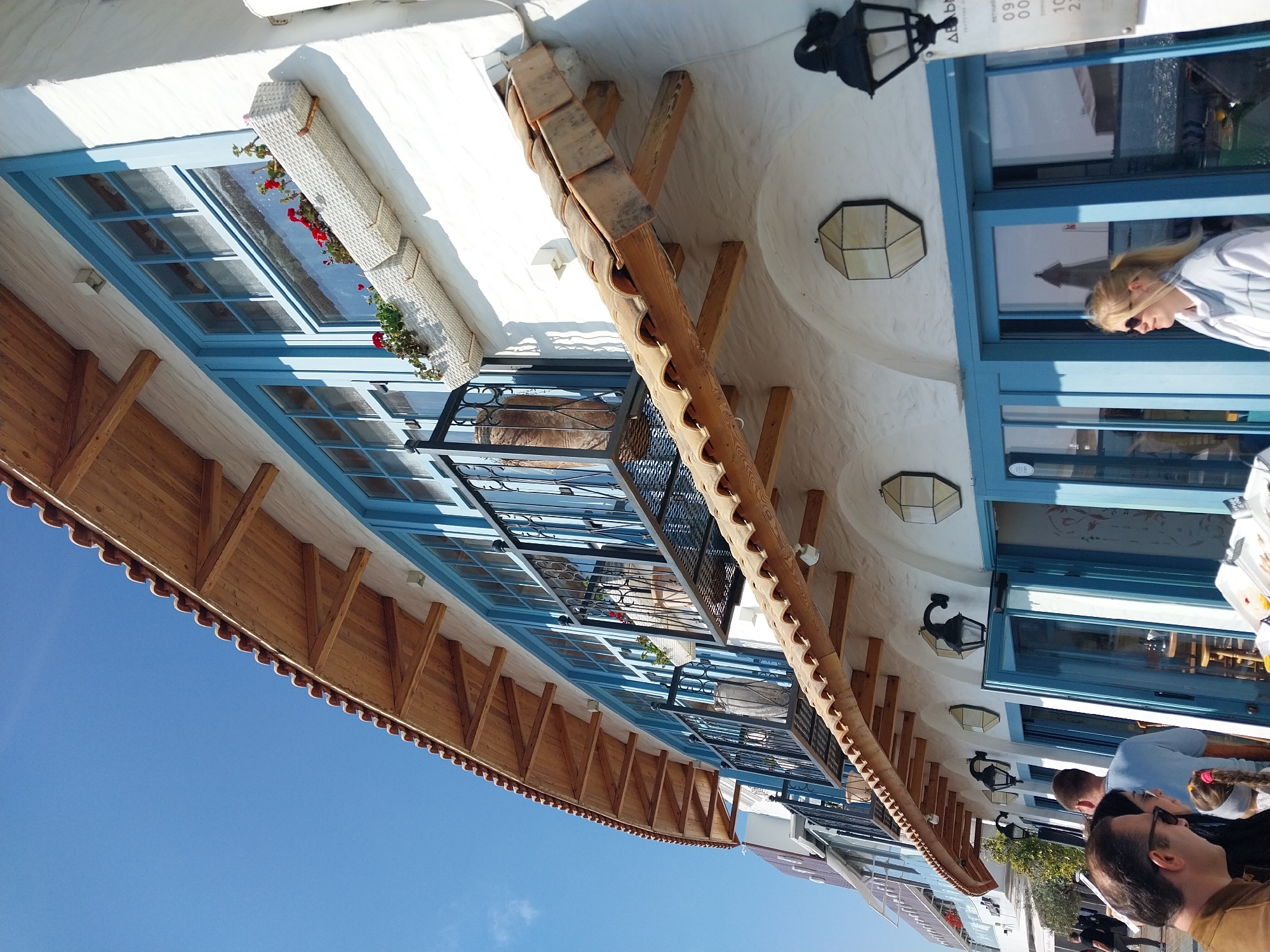 The image displays a sunny outdoor scene, likely a cafe or restaurant terrace, with the camera oriented vertically, placing the clear blue sky on the left and a building facade on the right.
The building features white stucco walls contrasting with vibrant blue window and door frames. Several sets of these blue-framed windows line the facade, some accompanied by decorative black iron railings, hinting at balconies. Below some windows, white wicker planter boxes hold red flowering plants. A prominent architectural feature is a long, horizontal wooden structure with terracotta tiles, possibly an awning or a decorative roof element, supported by a series of wooden beams. Hexagonal light fixtures and traditional black wall-mounted lamps are visible on the white walls.
Several people are present, indicating a social or dining activity. On the far right, a blonde woman wearing sunglasses and a light-colored top looks outward, her reflection visible in a window, which also reflects what appears to be water or a distant building. In the bottom right foreground, two men, one with dark hair and sunglasses, and another with shorter hair, are seen from behind, seated at a table alongside a child with a pink hair tie. Other figures are indistinct in the background.
The bright daylight and clear blue sky suggest a pleasant, sunny day. The architectural style, with white walls, blue frames, and terracotta, evokes a Mediterranean or coastal aesthetic. A white sign near the top right displays partial text in what appears to be Greek script ("ΔΕΛΦ...") and numbers ("09 00", "10 2..."), possibly indicating operating hours.