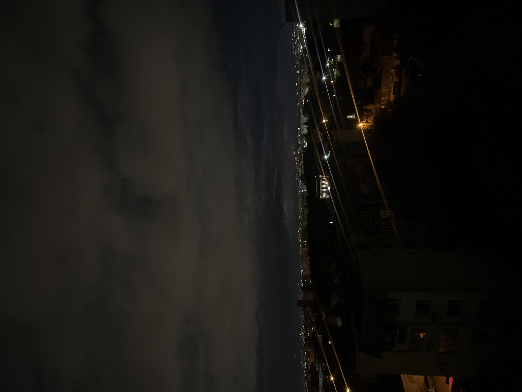 The image shows a nighttime view of Sofia, Bulgaria. In the distance, a city skyline is visible with numerous lights indicating buildings and streets. The sky is cloudy. The photograph appears to be taken from an elevated position, possibly a high floor of a building. Streetlights and the outlines of buildings closer to the camera are visible. The surrounding area immediately in front of the camera is dark.