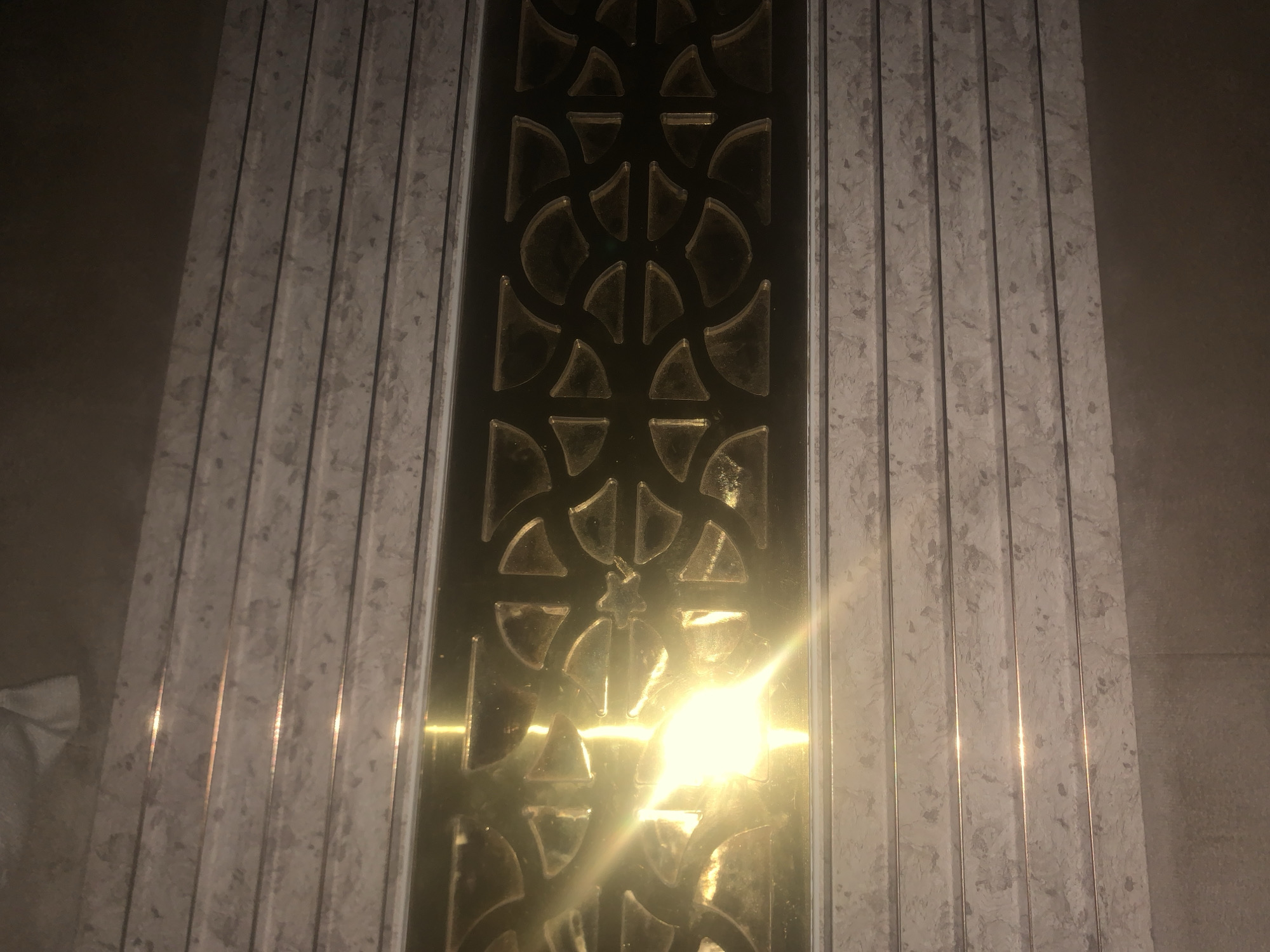 The image shows a close-up view of a decorative vertical element, possibly a panel or a column, featuring a central section with a golden, reflective metallic material adorned with a repeating geometric pattern. This pattern consists of curved and triangular shapes, suggesting a modern or art deco design. The central metallic section is flanked by two vertical panels that appear to be made of a light-colored material with a speckled or marbled texture, possibly stone or laminate. These flanking panels have a fluted or ribbed surface, with each rib separated by a dark, recessed line, creating a sense of depth and verticality. A bright light source, possibly a flash or sunlight, is reflecting strongly off the golden central section, creating a lens flare effect and washing out some of the details in that area. The background is dimly lit and appears to be a plain, neutral surface, possibly a wall. The image is focused on the decorative element and does not provide any context for its location in Jalingo, Nigeria. There are no people, animals, or specific actions depicted. The scene is static and focuses solely on the architectural or decorative detail.