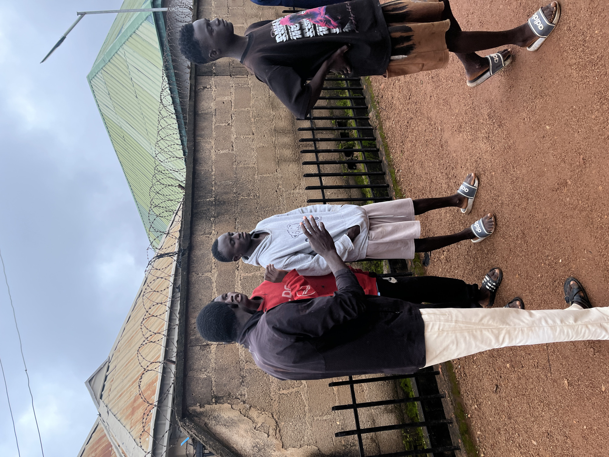 The image captures a group of young men in Barakin Akawo, Nigeria, standing near a brick wall topped with barbed wire. The sky is overcast. In the foreground, four men are visible. One man on the far left, wearing a dark jacket and light-colored pants, is engaged in conversation with two other men. One of these men, in a red hoodie, is looking towards the speaker, while the other, in a grey hooded sweatshirt and light shorts, gestures with his hands. A fourth man on the right, wearing a dark t-shirt and light brown shorts, stands with his hands on his hips, looking to the side. The ground is covered in reddish-brown gravel. Behind the men, a black metal fence runs along the base of the brick wall. Two rooftops are visible above the wall: one appears to be corrugated iron with some rust, and the other is a green, ribbed metal roof. A street lamp extends from behind the green roof.