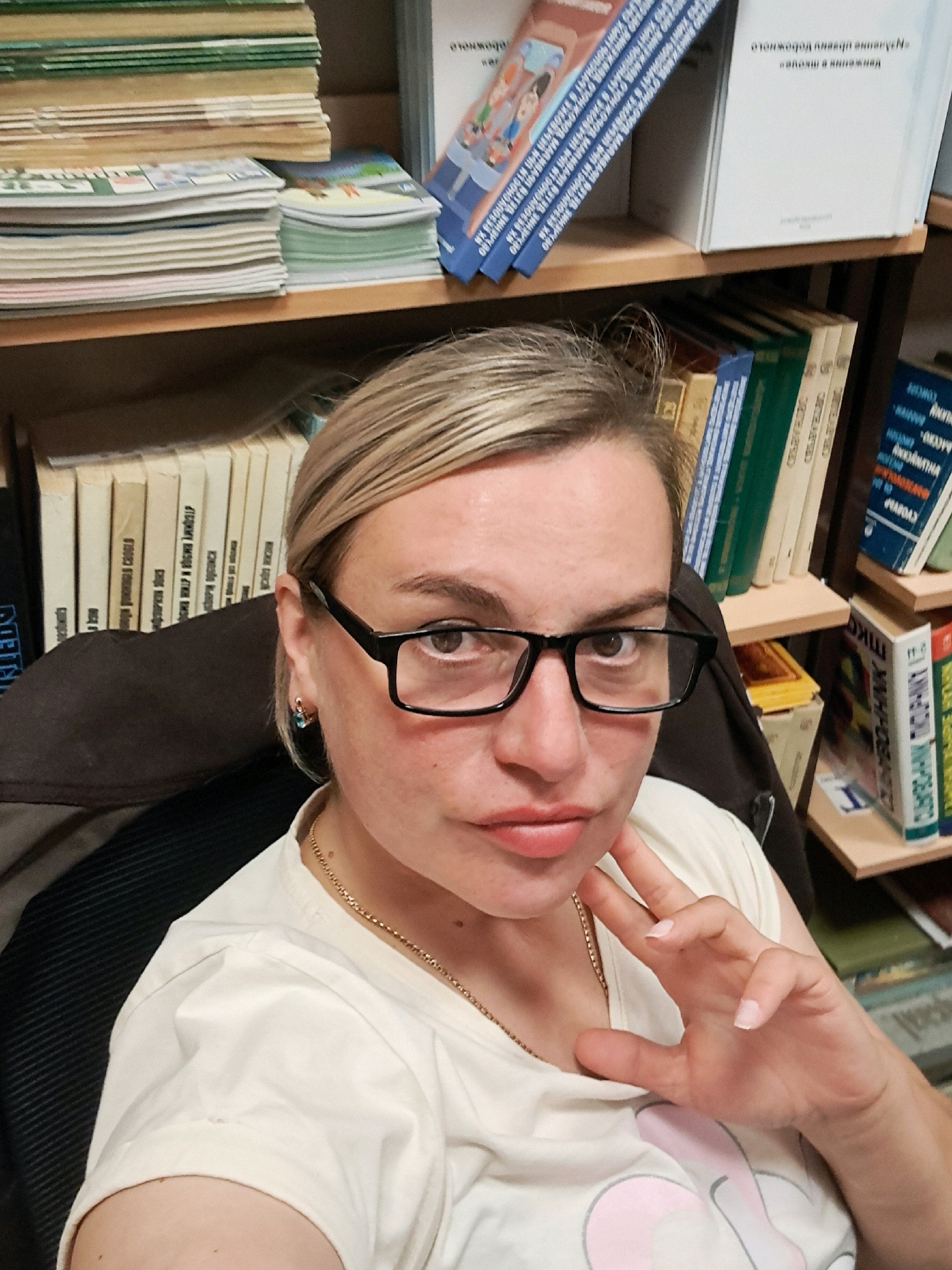 The image shows a close-up, selfie-style photograph of a woman with blonde hair and glasses, sitting in front of bookshelves. The woman is wearing a light-colored t-shirt and a gold necklace. Her right hand is resting on her chin, and her left hand is visible in the foreground. The bookshelves behind her are filled with various books. The overall environment appears to be a library or study. The image does not contain any specific elements that would allow for definitive identification of the location within the city of Elektrostal, Russia.
