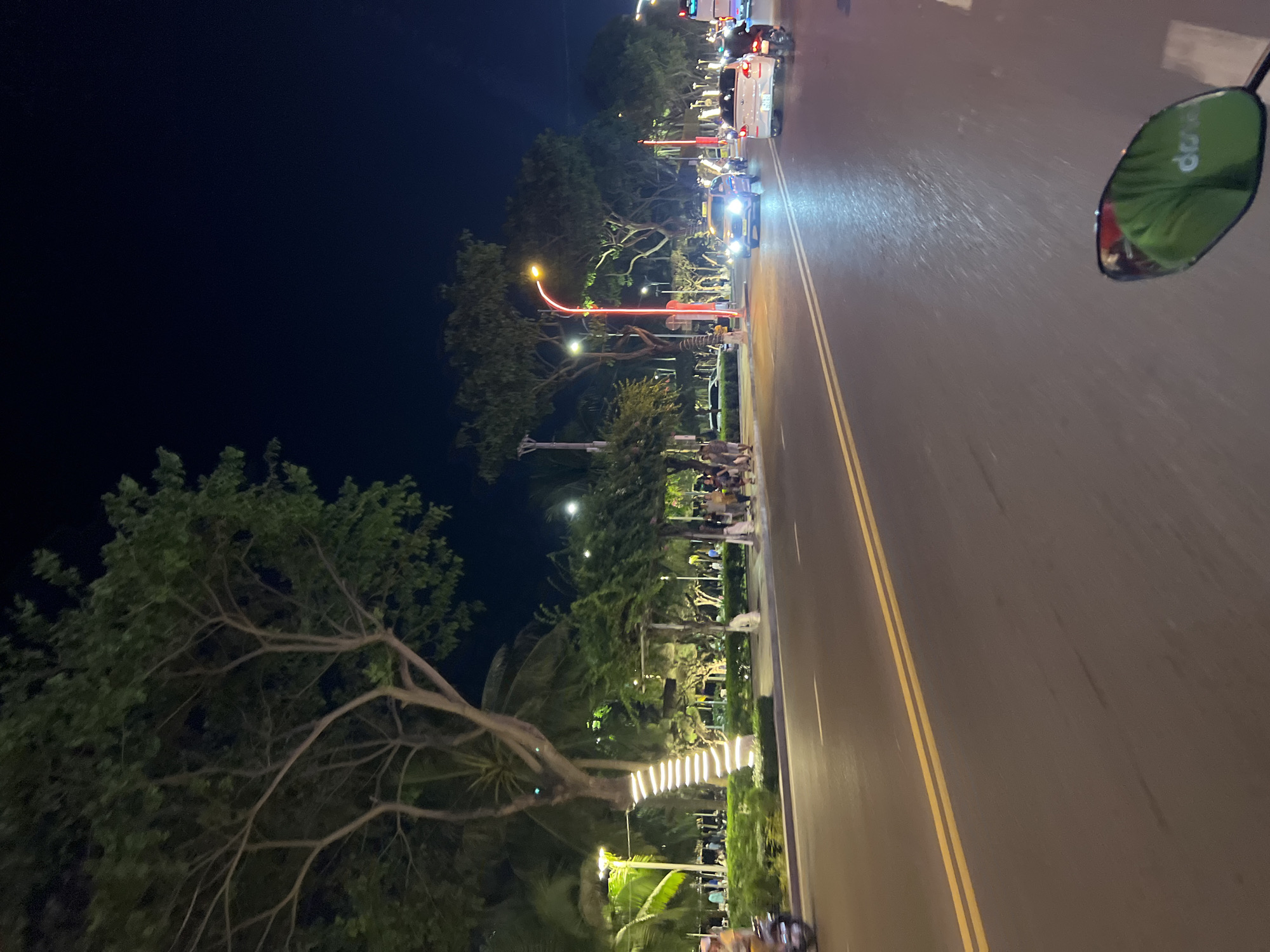 The image depicts a nighttime scene on a road in Nha Trang, Vietnam. The view is from the perspective of someone riding a motorcycle, evident from the side mirror in the top right corner. The road is lit by streetlights and headlights from oncoming traffic. Along the side of the road, there is a park area with trees, some decorated with lights. People are visible along the park, suggesting a pedestrian area or gathering space. In the distance, more vehicles and streetlights suggest a busy urban area. The general atmosphere is that of a lively evening scene in a coastal city.