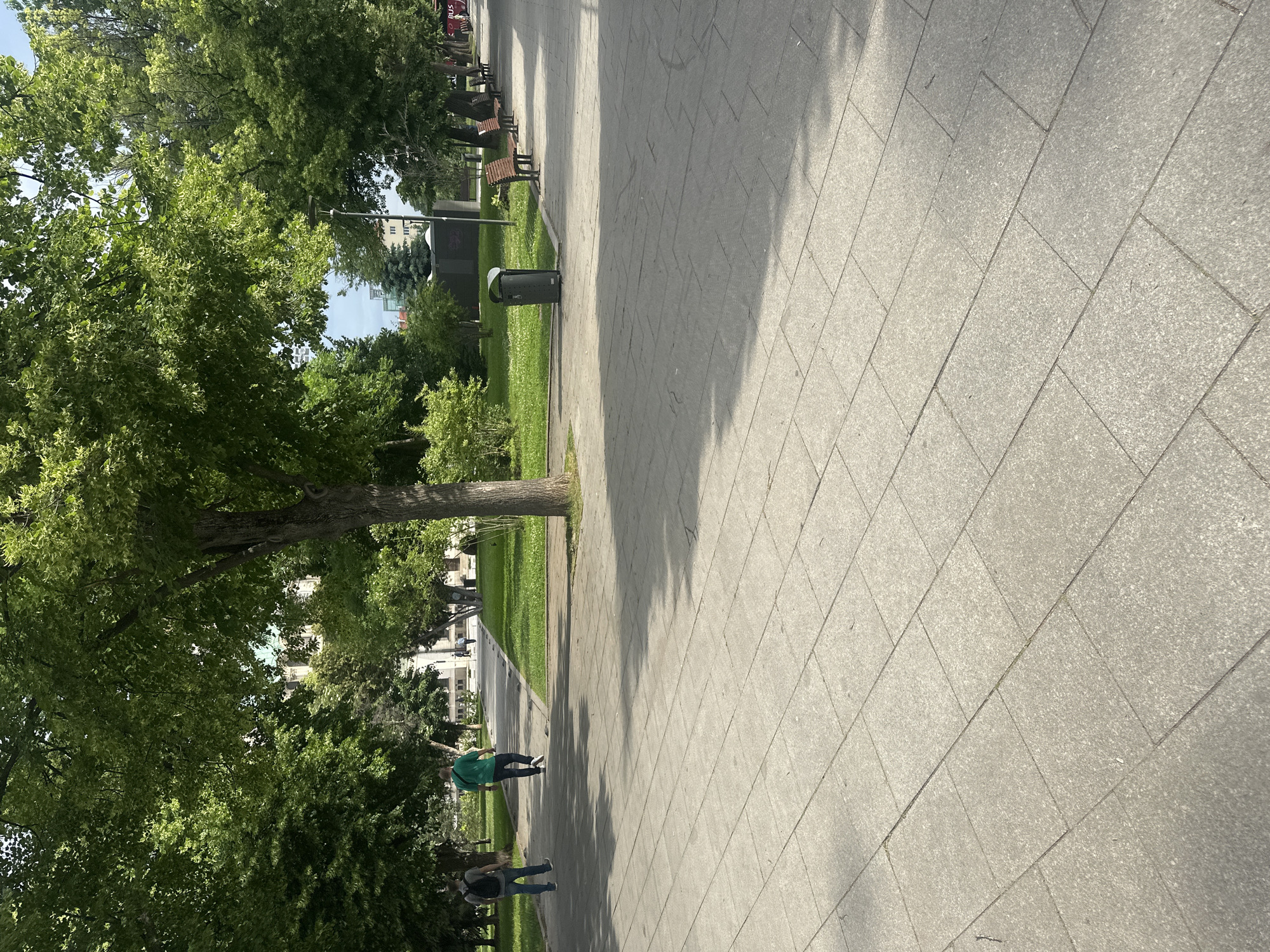 This photo, taken in Sofia, Bulgaria, depicts a tree-lined paved walkway with a grassy area on the left. A trash can sits next to a park bench. Two pedestrians are walking in the distance. The pavement is made of large grey tiles. The day appears sunny, with shadows visible on the ground.