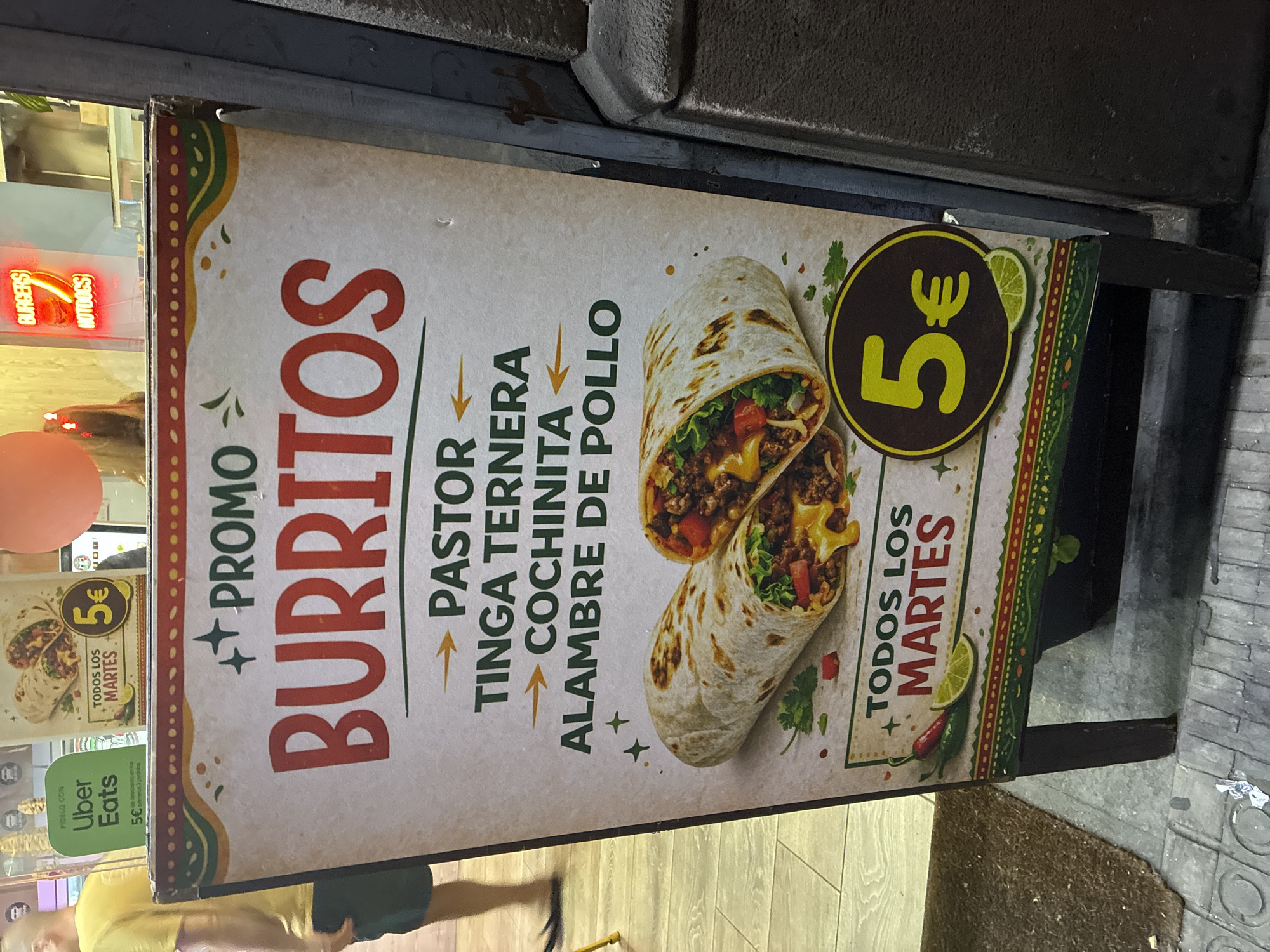 A vertical, rectangular A-frame sandwich board sign, dark-framed, is positioned on an urban sidewalk or entranceway in Barcelona, Spain. The sign, predominantly white with a decorative red, green, and yellow patterned border, prominently advertises "PROMO BURRITOS" with "BURRITOS" in large red capital letters. Below this, four burrito filling options are listed in green text with yellow directional arrows: "PASTOR," "TINGA TERNERA," "COCHINITA," and "ALAMBRE DE POLLO." The lower half of the sign displays a graphic of two burritos, one whole and one cut in half revealing its interior of ground meat, melted cheese, lettuce, and diced tomatoes. Illustrations of fresh cilantro, red and green chili peppers, and lime wedges are scattered around the burritos. In the bottom right corner, a prominent yellow circle contains the text "5€," and below it, a lime wedge is depicted. To the left of the yellow circle, the text "TODOS LOS MARTES" (All Tuesdays) is displayed in green, with "MARTES" in red.
In the background, a shop window reveals parts of an establishment's interior and additional signage. To the upper left, a green sign for "Uber Eats" is visible, stating "PIDELO CON" (Order with) and offering "5€ de descuento en tus 2 primeros 2 pedidos" (5€ discount on your first 2 orders). Another smaller sign, similar in design to the main burrito promotion, is also visible. To the upper right, a red neon sign inside the shop spells out "BURGERS HOTDOGS." An orange, spherical object, likely a balloon, is partially visible in the top right corner of the frame.
A male figure, with light skin and wearing a yellow t-shirt and dark shorts, is partially visible on the left side of the frame, walking from right to left, away from the camera. His left leg and foot, clad in a dark flip-flop or sandal, are clearly visible.
The ground in the foreground consists of a dark brown, textured mat on the left. The central area features light-colored square floor tiles with visible grout lines, suggesting an indoor or covered outdoor area. A dark, vertical, possibly concrete or stone structural pillar or wall runs along the right edge of the image. The scene is artificially lit by ambient light from the shop and neon signs. The use of the Euro currency symbol (€) and Spanish language on the signs confirms the location within a Spanish-speaking European city, specifically Barcelona as per instruction.