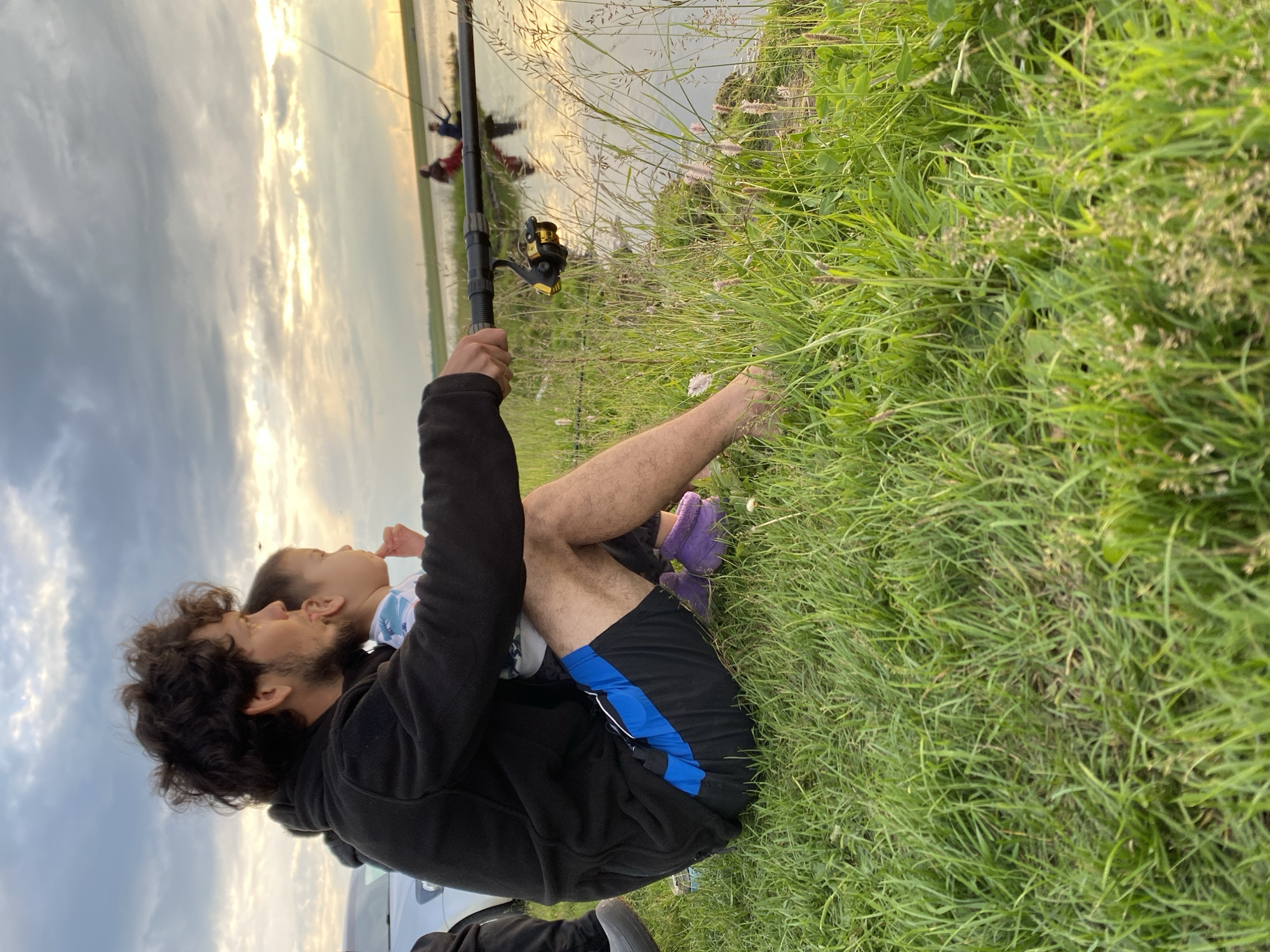 A man and a child are sitting on the grass by the edge of a body of water, fishing. The man has curly dark hair and a beard and is wearing a black hooded jacket and blue and black shorts. He is holding a fishing rod with a child sitting on his lap and looking in the same direction. The child is wearing a blue and white patterned shirt and purple slippers. The water reflects the cloudy sky, which is illuminated by the setting sun. In the background, two other figures are visible in the water, also fishing. The area around them is covered in green grass and some wildflowers. The scene appears to be taking place in the village of Syryshbashevo, Russia.