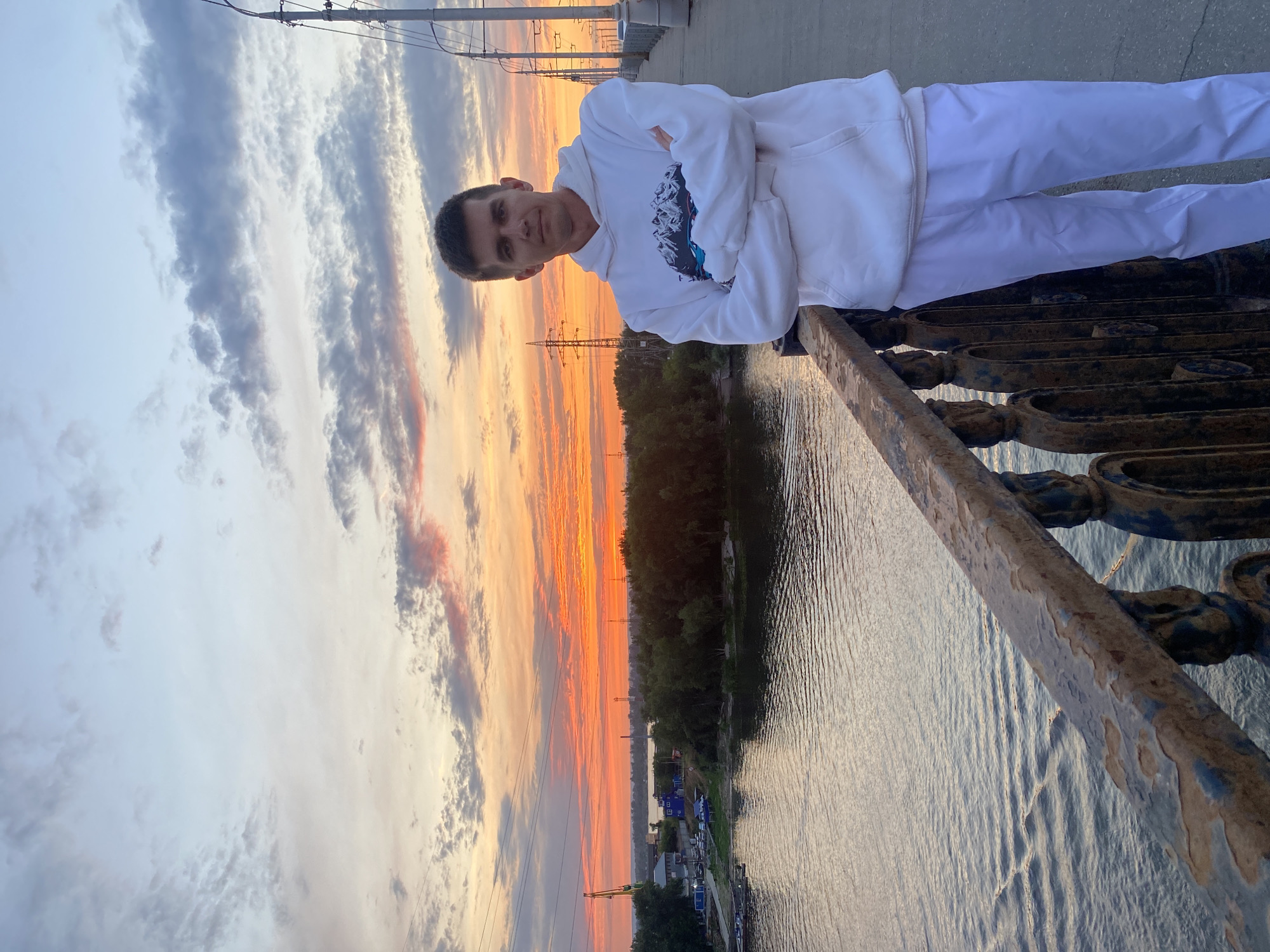 A young man stands on a bridge over a river at sunset in Novokuybyshevsk, Russia. He is wearing a white hoodie with a graphic of mountains and white pants. His arms are crossed. The sky is filled with vibrant orange and pink clouds, reflecting on the water. In the background, there are trees, industrial buildings, and power lines. The bridge has a weathered metal railing in the foreground.