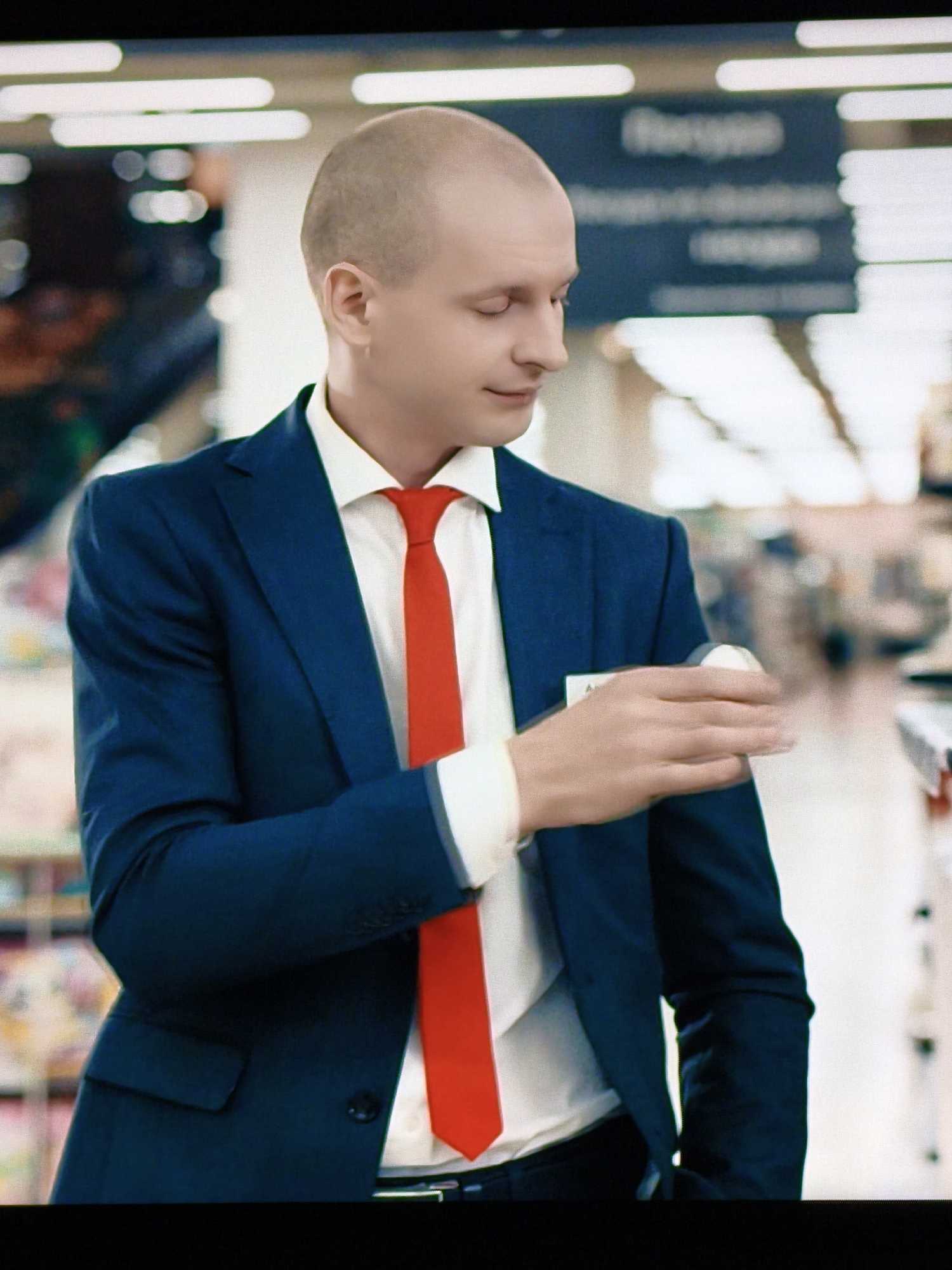 A bald Caucasian man in a dark blue suit, white shirt, and red tie is shown from the chest up. He is wearing a nametag on his left lapel. The man is looking down and to his right, with his right hand near his chest, holding a small white object. The background is blurred and appears to be an indoor environment, possibly a store or office, with lights and shelves visible. There is no indication of the location being Vladivostok, Russia.