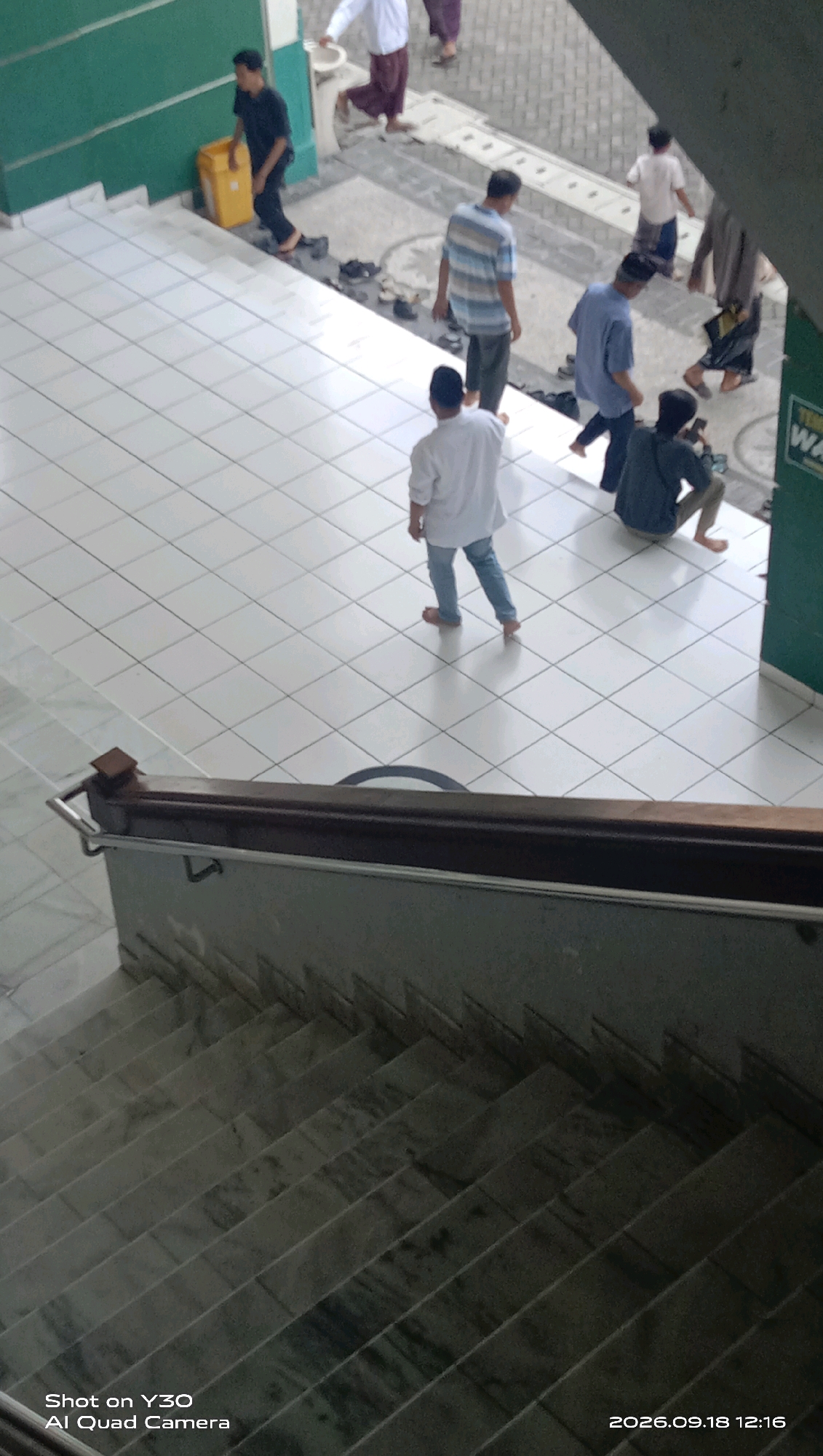 The image depicts an indoor scene with a group of individuals present. In the foreground, a marble staircase is visible, descending into shadow. A handrail made of dark wood and metal runs along the side of the staircase. Above the staircase, a tiled floor stretches out, with a diamond pattern formed by the white tiles. Several people are scattered across this tiled area. One individual, wearing a white long-sleeved shirt and blue jeans, stands with their back to the camera, facing away from the stairs. To the right of this person, another individual in a dark long-sleeved shirt and dark pants sits on the floor, looking at a mobile device. Further back, a person in a blue long-sleeved shirt and dark pants walks away from the camera, and another person in a striped blue and white long-sleeved shirt and grey pants also walks away from the camera. To the left, a person in a black long-sleeved shirt and dark pants stands near a yellow container. In the background, several other individuals are partially visible, some standing and some walking. The walls are painted in green and white. The image is dated "2026.09.18 12:16". The scene appears to be located within a public building, possibly a place of worship or a community center, given the attire of some individuals and the presence of a yellow container which could be for donations or waste. The overall lighting suggests it is daytime.