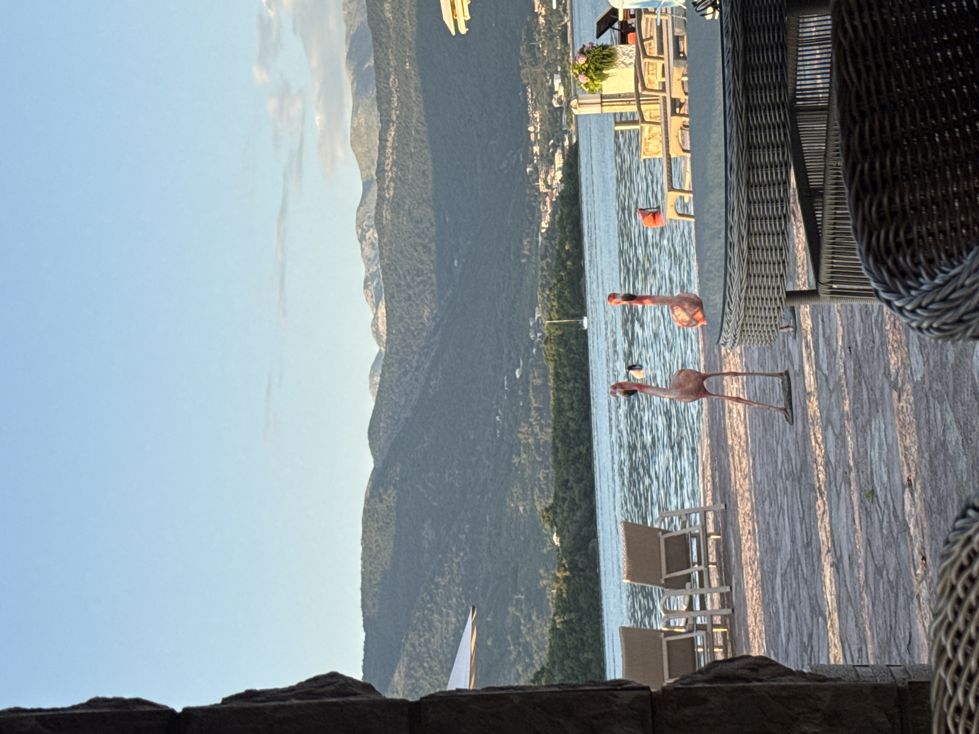 The image shows a view from a patio, presumably in Bogišići, Montenegro. The foreground is comprised of a low stone wall and woven outdoor furniture. Beyond the patio is a lake and forested mountains. There are pink flamingo statues on the patio. In the lake, a white sailboat can be seen. Other patio furniture, tables, and potted flowers are visible closer to the water's edge. The sky is light blue.