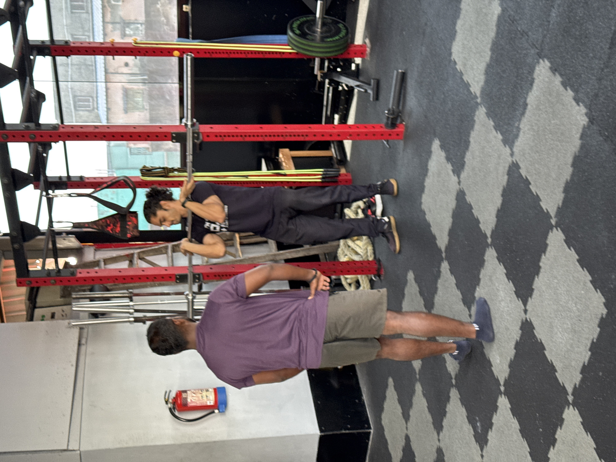 A fitness gym in Kolkata, India, is set up with a squat rack loaded with a weight plate. Two men are in the gym: one stands with his back facing the camera wearing a purple t-shirt and tan shorts. The other man is using resistance bands attached to the rack. The room has interlocking foam mats with a diamond pattern. A fire extinguisher is attached to the wall.
