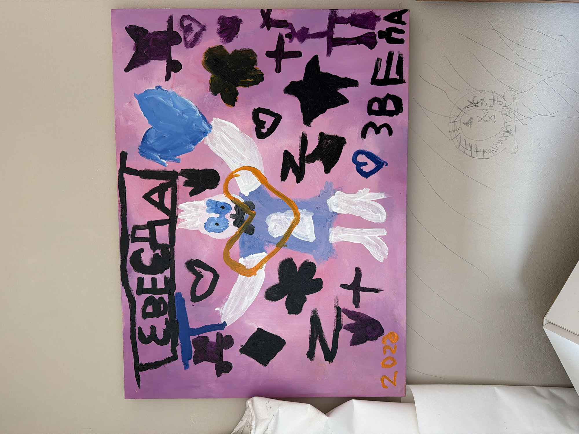 A painting is displayed on a wall. The painting is done on a rectangular canvas with a pink and purple gradient background. At the top of the painting, in black, are the Cyrillic letters "ЛЕБЕДЯ" which translates to "SWAN". Below this text, various colorful shapes and figures are depicted. There is a large blue heart, a drawing of a figure with white limbs and a blue and purple torso, outlined in black. An orange heart shape encircles the head and upper torso of this figure. There are also black shapes resembling hearts, a diamond, a star, and a flower-like form. To the lower left of the painting, the year "2023" is written in orange. To the lower right of the painting, the Cyrillic letters "ЗВЕЗДА" which translates to "STAR" are written in black. The painting appears to be a child's artwork. The wall behind the painting is a light beige color with faint, light gray diagonal lines. To the left of the painting, a roll of white paper is partially visible. Below the painting and to the right, there is a circular drawing on the wall, possibly made with a pencil, containing some indistinct shapes and what might be letters. No specific location in the city of Ufa, Russia can be determined from the image.