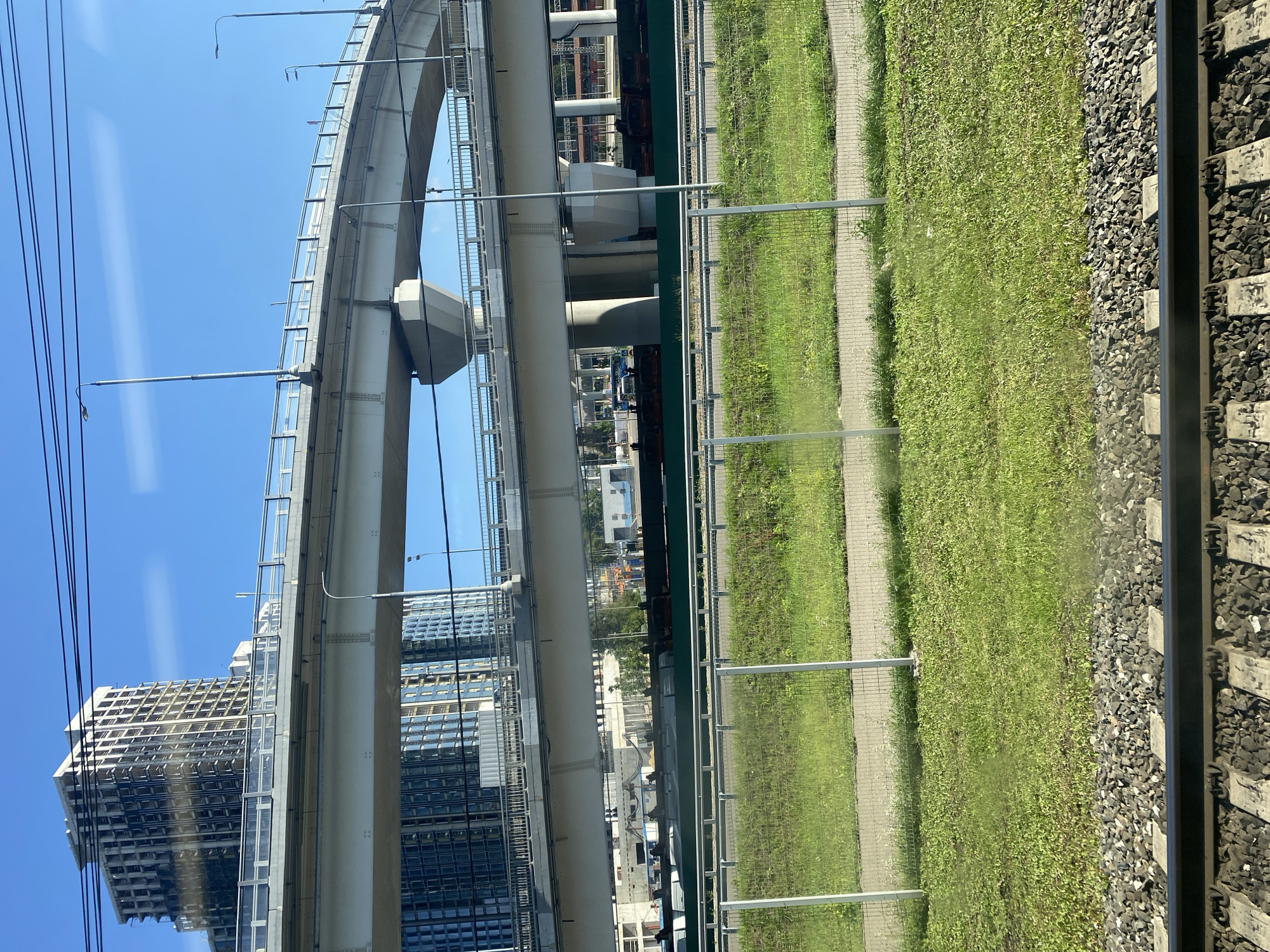 The image displays a modern cityscape with multi-level overpasses and tall buildings under construction. The foreground shows a section of railroad tracks, indicating the photo was likely taken from a moving train. Beyond the tracks, a strip of green grass is visible, bordered by a chain-link fence. The overpasses, made of concrete, dominate the mid-ground, with one appearing to be a significant highway interchange curving into the distance. The background features two large, contemporary buildings, one of which is clearly in an unfinished state, revealing its skeletal structure and exposed framework. The sky is a clear, bright blue, suggesting a sunny day. The overall scene suggests an urban environment undergoing development.