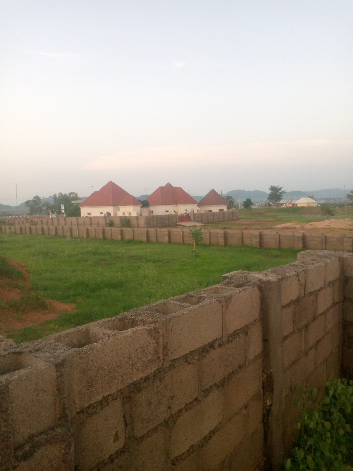 The image depicts an overcast sky over a residential construction site in Jalingo, Nigeria. In the foreground, a wall constructed from concrete blocks is visible. Beyond this wall, a grassy field stretches towards a series of smaller, uniform walls also made of concrete blocks. Further in the background, three houses with distinctive red, pyramid-shaped roofs are situated behind the block walls. These houses appear to be under construction or recently completed. The environment is mostly green with patches of bare earth. In the distance, hills and more buildings can be observed under the hazy sky. There are no people visible in the image.