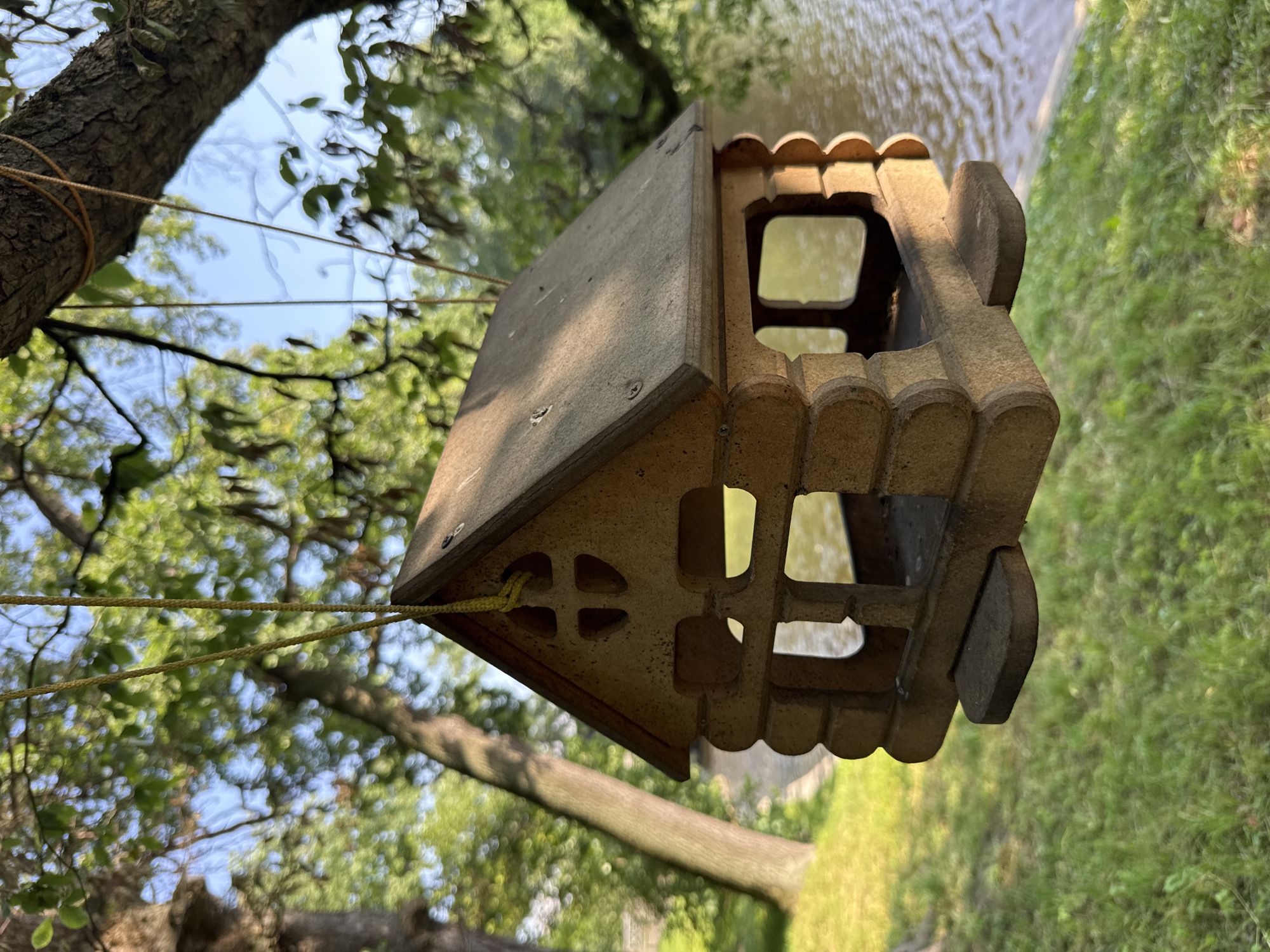 This media file shows a wooden birdhouse hanging from a tree branch by yellow rope. The birdhouse is designed to resemble a small house with multiple openings and decorative trim. In the background, out-of-focus trees and greenery are visible, along with a body of water, suggesting an outdoor setting in a park or natural area. The lighting suggests it is daytime, possibly late afternoon due to the warm tones. The scene appears peaceful and serene. No people are visible, and there is no visible text. The context provided indicates the location is Saint Petersburg, Russia.