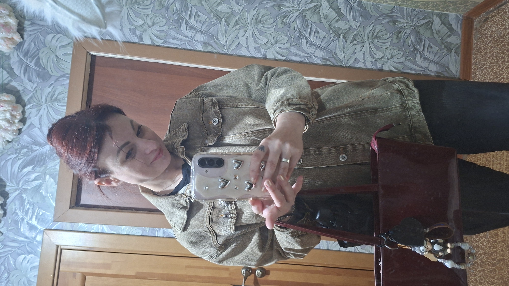 A woman with red hair is taking a selfie in a mirror. She is wearing a tan denim jacket and black pants. She is holding a white phone with heart-shaped decorations on the case. A burgundy purse is also visible. The background shows a bedroom with a patterned wallpaper and a wooden dresser. The location is Забалоцкі сельскі Савет, Belarus.