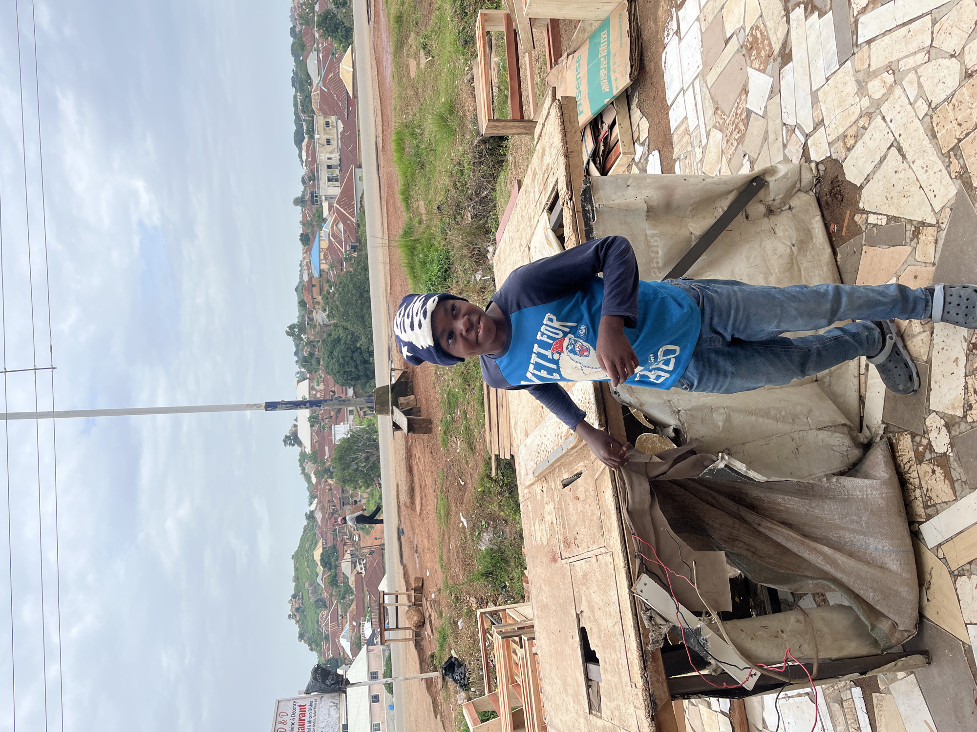 A young Black boy wearing a blue long-sleeved shirt with "Yeti for Prez" printed on it and a black and white patterned beanie stands in front of a makeshift stall in Jos, Nigeria. The stall is constructed from rough wood and covered with torn, tan tarp. Exposed wires are visible, suggesting it may have been used for electrical equipment. The ground is covered in a mosaic of broken tiles and dirt. In the background, a sprawling cityscape of red-roofed buildings stretches across rolling hills under an overcast sky, with a billboard for "D&D Divine & Decency Restaurant" visible to the left. Other construction materials, including wooden crates and what appear to be disassembled furniture, are scattered around the immediate area. A person walks in the distance on a road winding through the city. The overall scene depicts a somewhat unkempt, possibly developing urban environment.