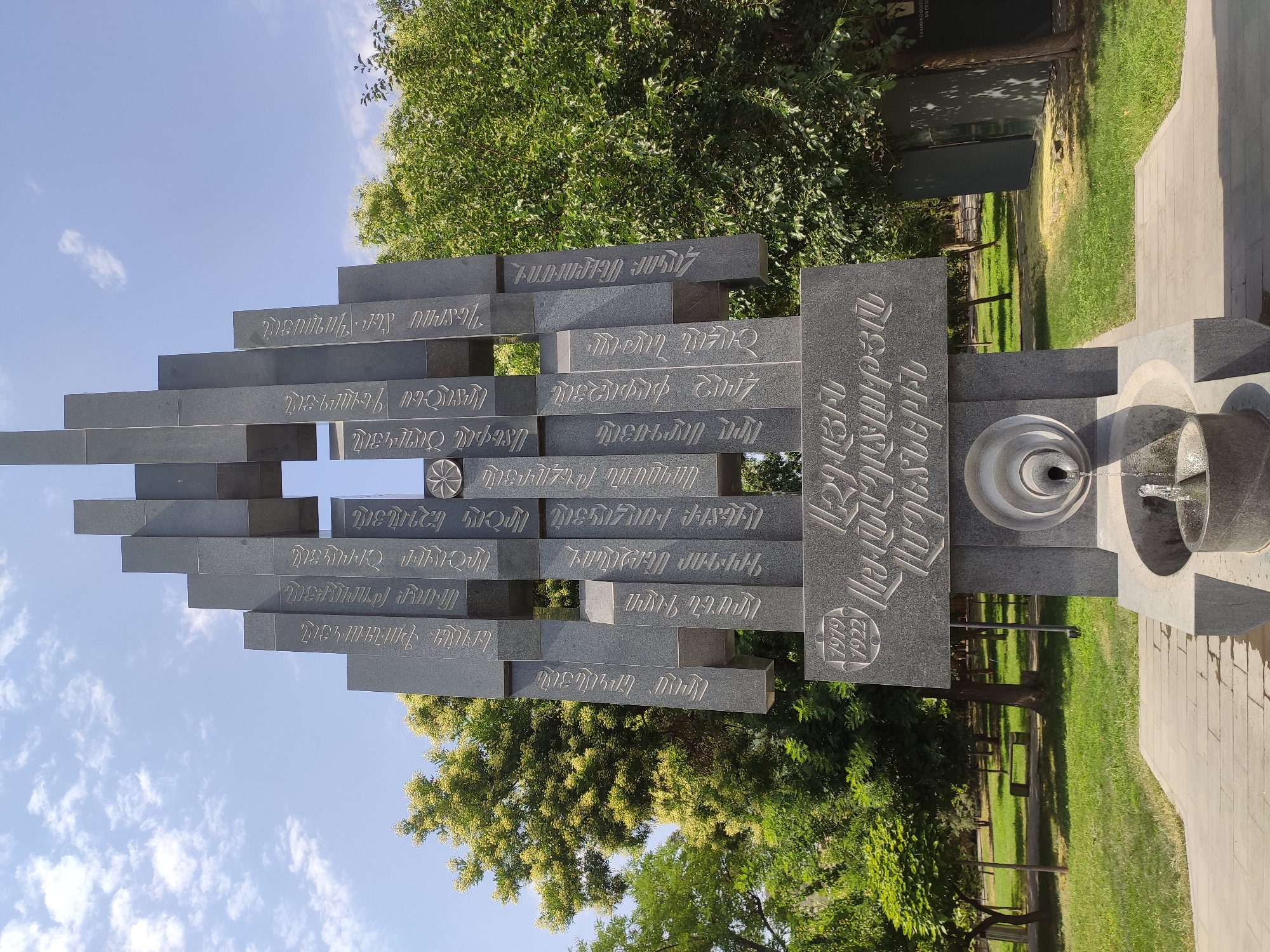 This is a low-angle, medium shot of a monument in Yerevan, Armenia, on a clear, sunny day. The monument is made of dark grey stone and features several vertical rectangular slabs arranged in an abstract, staggered formation. Inscribed on the slabs are Armenian inscriptions and what appears to be a floral symbol. The monument has a fountain at its base, with water flowing into a stone basin. The monument is set in a park with green grass, trees, and a paved walkway in the foreground. The sky is blue with scattered white clouds.