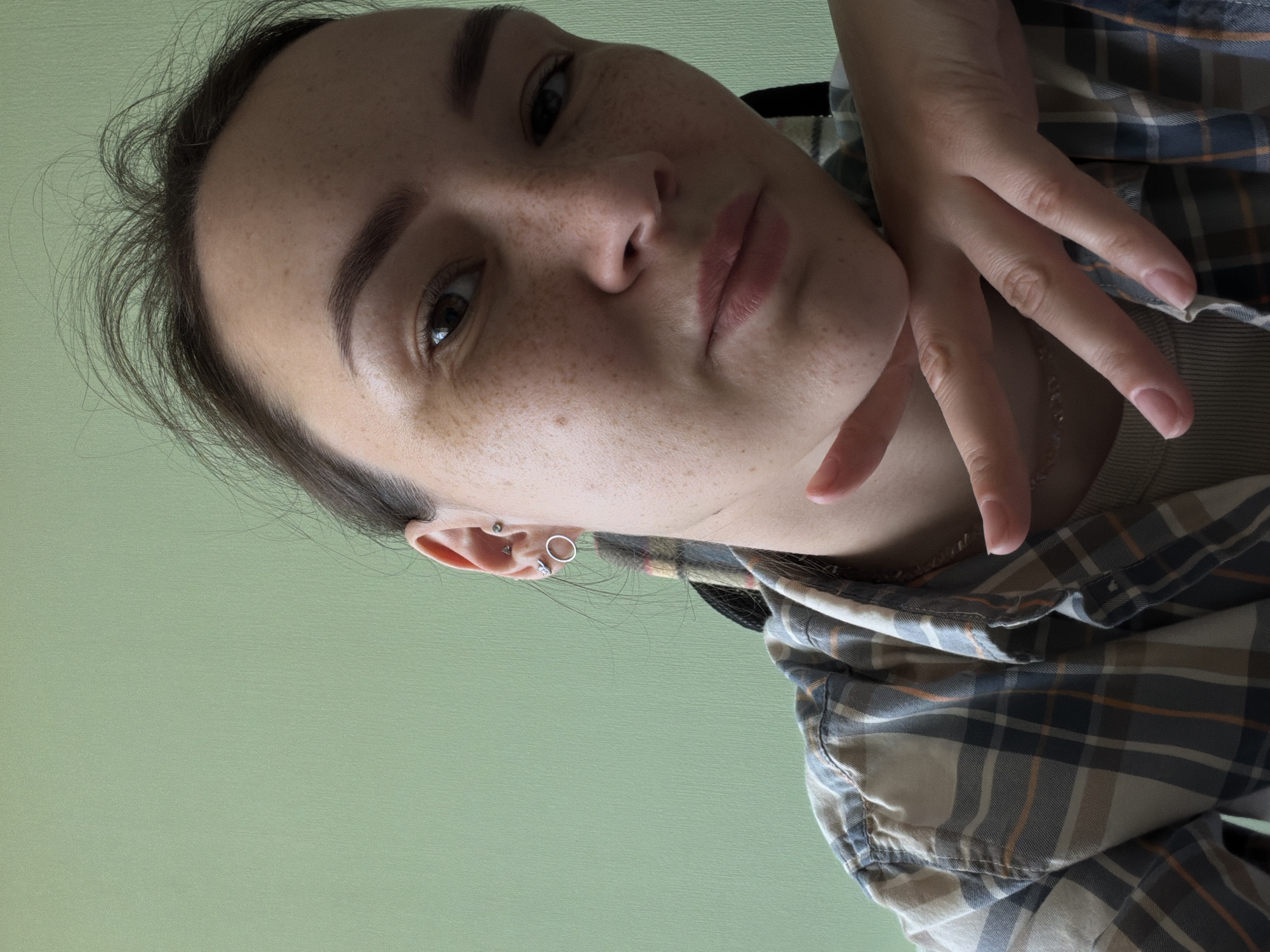 A young woman with dark hair, freckles, and dark eyes is shown in a close-up, selfie-style photograph. She is wearing a plaid shirt over a tan ribbed tank top and has a delicate necklace. Her right hand is resting on her chin and cheek. She has multiple ear piercings, including a circular stud and a small hoop. The background is a plain, light green wall. The image appears to have been taken indoors. There is no information in the image to suggest a location in the city of Chaikovsky, Russia.
