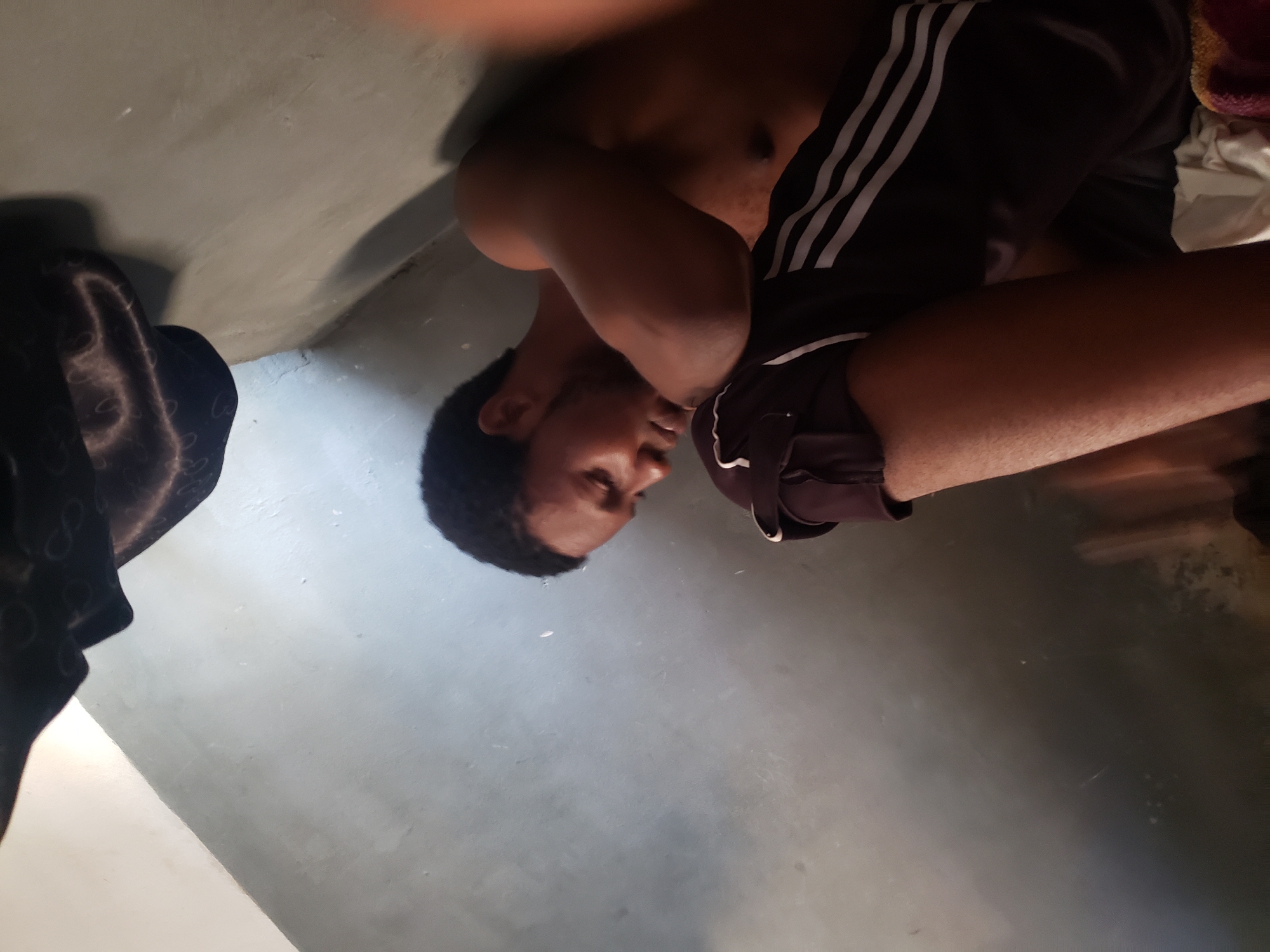 An adult male is depicted in an indoor setting, lying down or leaning against a light-colored, plain surface. The image is significantly tilted, making it appear as if he is positioned in a corner where a wall meets a floor, both made of a textured, possibly concrete or painted plaster material. The man has short, dark hair and a neutral expression, looking slightly downwards. He appears to be shirtless, showcasing his torso, and is wearing dark athletic shorts with three distinct white stripes on the side. To the left, a dark, patterned fabric, possibly a blanket or piece of clothing, is visible. Another lighter-colored fabric, which could be a towel or cloth, is present near his right hip. The lighting is dim and uneven, casting soft shadows across the surfaces. There are no discernible specific activities, visible text, or strong location cues within the image itself.